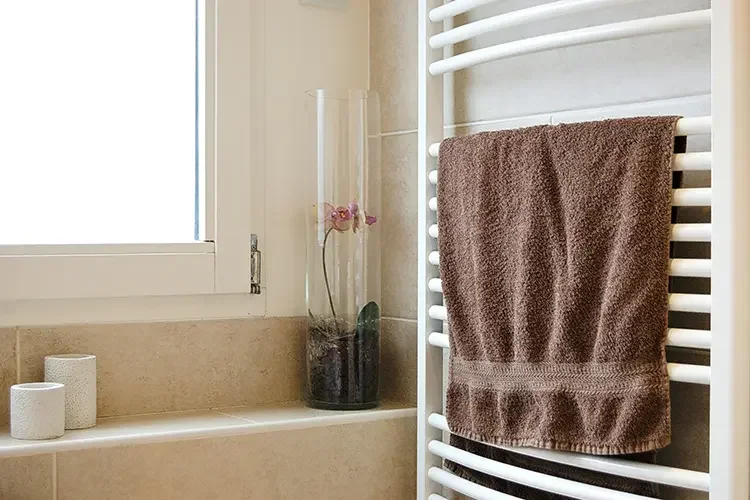 A fluffy brown bath towel hanging flat to dry on a white heated towel rack next to a window in a modern, minimalist bathroom.