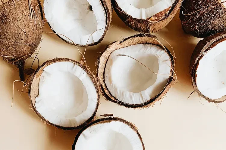 Top-down view of several fresh, halved coconuts with clean white flesh scattered on a warm beige background, representing the natural ingredient.