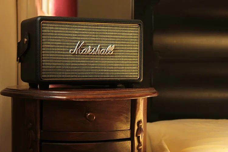 A classic, sophisticated Marshall vintage Bluetooth speaker rests on an elegant wooden nightstand. Warm, ambient lighting softly highlights the iconic gold logo and textured grille, creating a cozy and peaceful atmosphere in the bedroom.