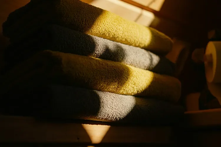 Stack of folded gray and mustard-yellow bath towels lit by warm afternoon sunlight.