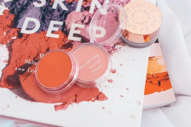 Two shades of peach and coral powder blush containers placed on top of a beauty magazine, showcasing professional makeup products for warm skin tones.