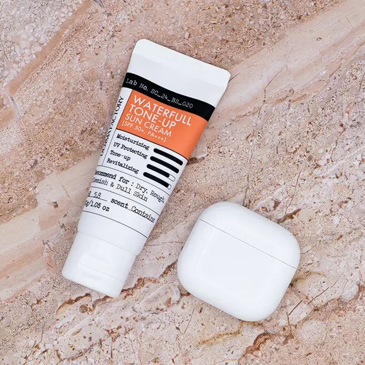 Derma Factory Waterfull Tone-Up Sun Cream tube next to white AirPods on a luxury beige marble background, featured on TheWellnessExaminer.