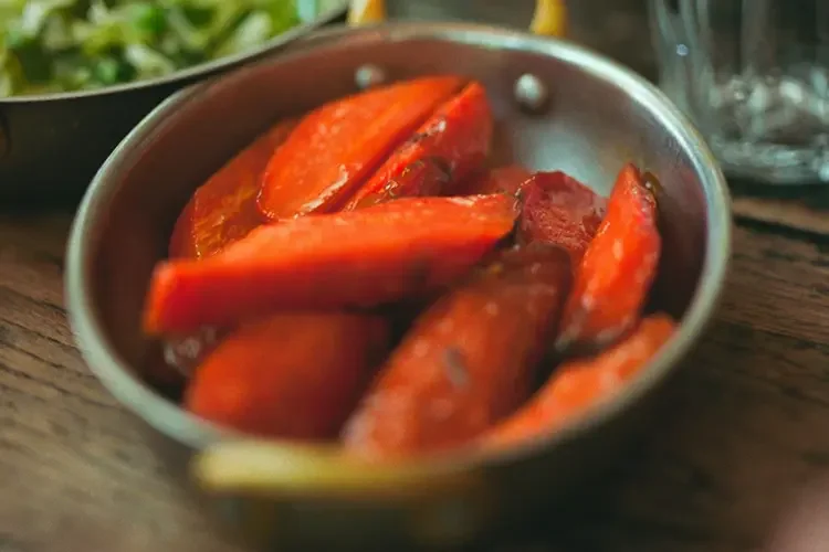 Glazed, cooked carrot wedges in a small metal pan on a wooden table, with a bowl of greens and a water glass at the edge of the frame.