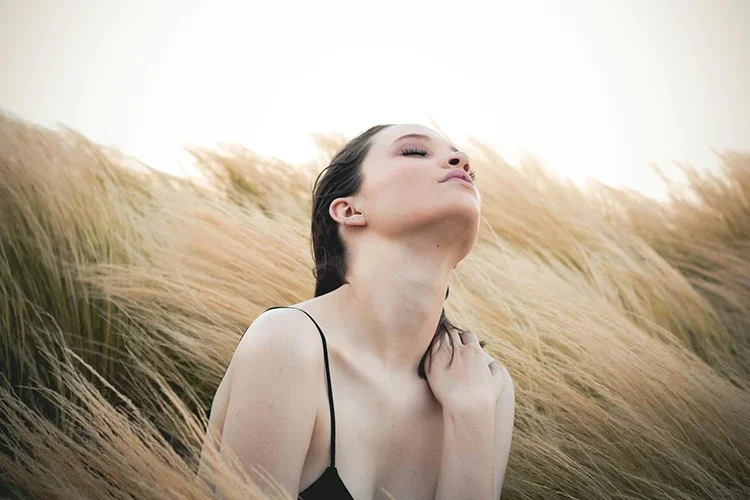 A serene woman with closed eyes tilting her head back and gently touching her shoulder in a field of tall grass, illustrating mindful deep breathing and nervous system relaxation.
