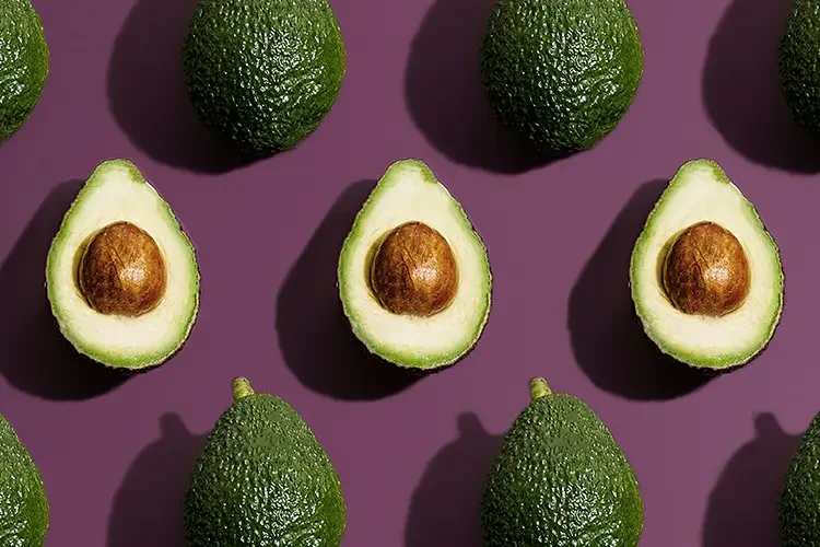 A high-angle pattern shot of halved avocados with pits on a purple background.