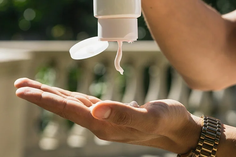 Close-up of a person squeezing lotion from a tube container into their hand under sunlight.