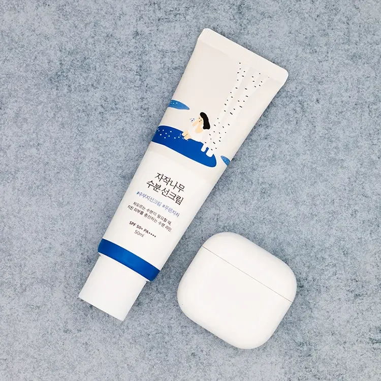 A square crop top-down view of ROUND LAB Birch Juice Moisturizing Sunscreen tube next to a white AirPods case on a textured blue stone surface for size comparison, featured on TheWellnessExaminer.