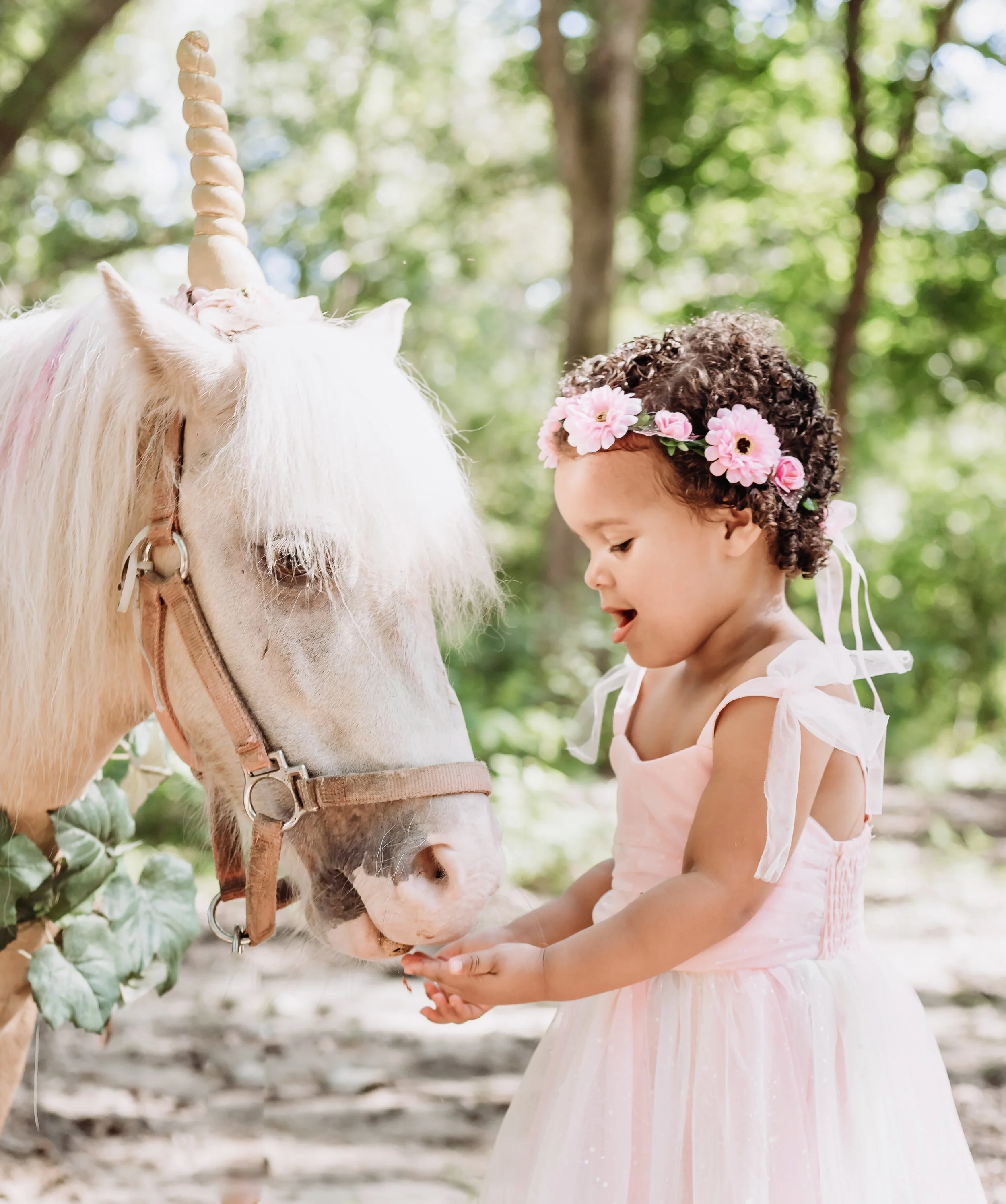 A young girl with curly hair wearing a pink dress and a flower crown is standing outdoors in a wooded area, gently holding a white unicorn.