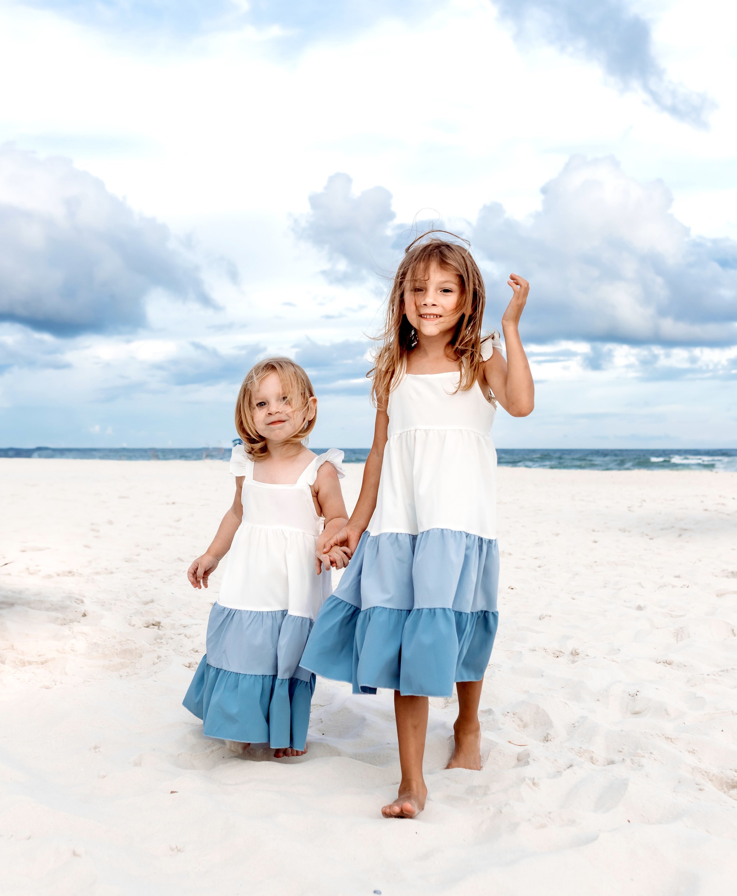 Two young girls in white and blue dresses walking on a sandy beach, holding hands, with the ocean and cloudy sky in the background.