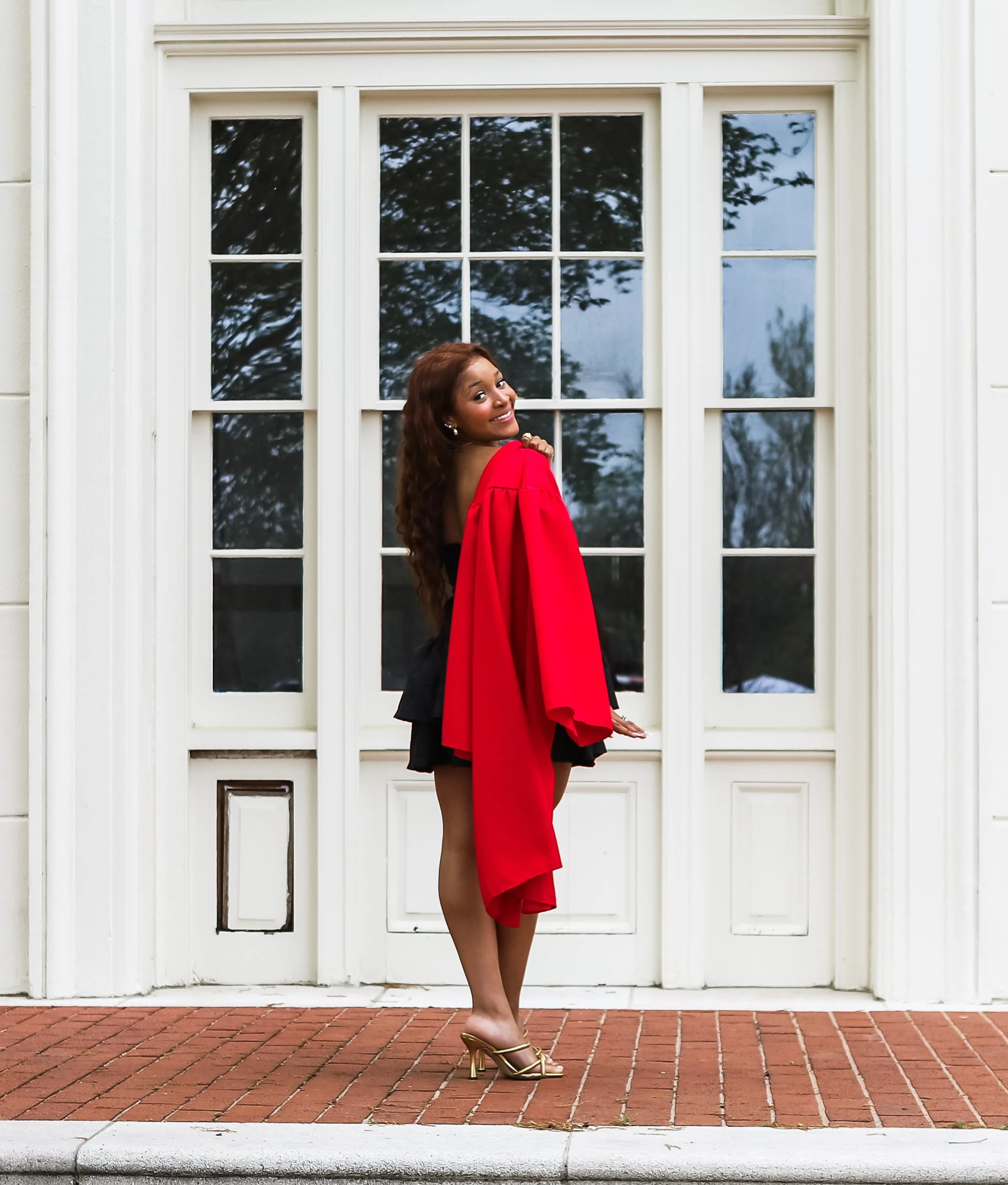 A woman in a black dress with a red graduation gown and gold heels stands in front of a white building with large windows, smiling at the camera.