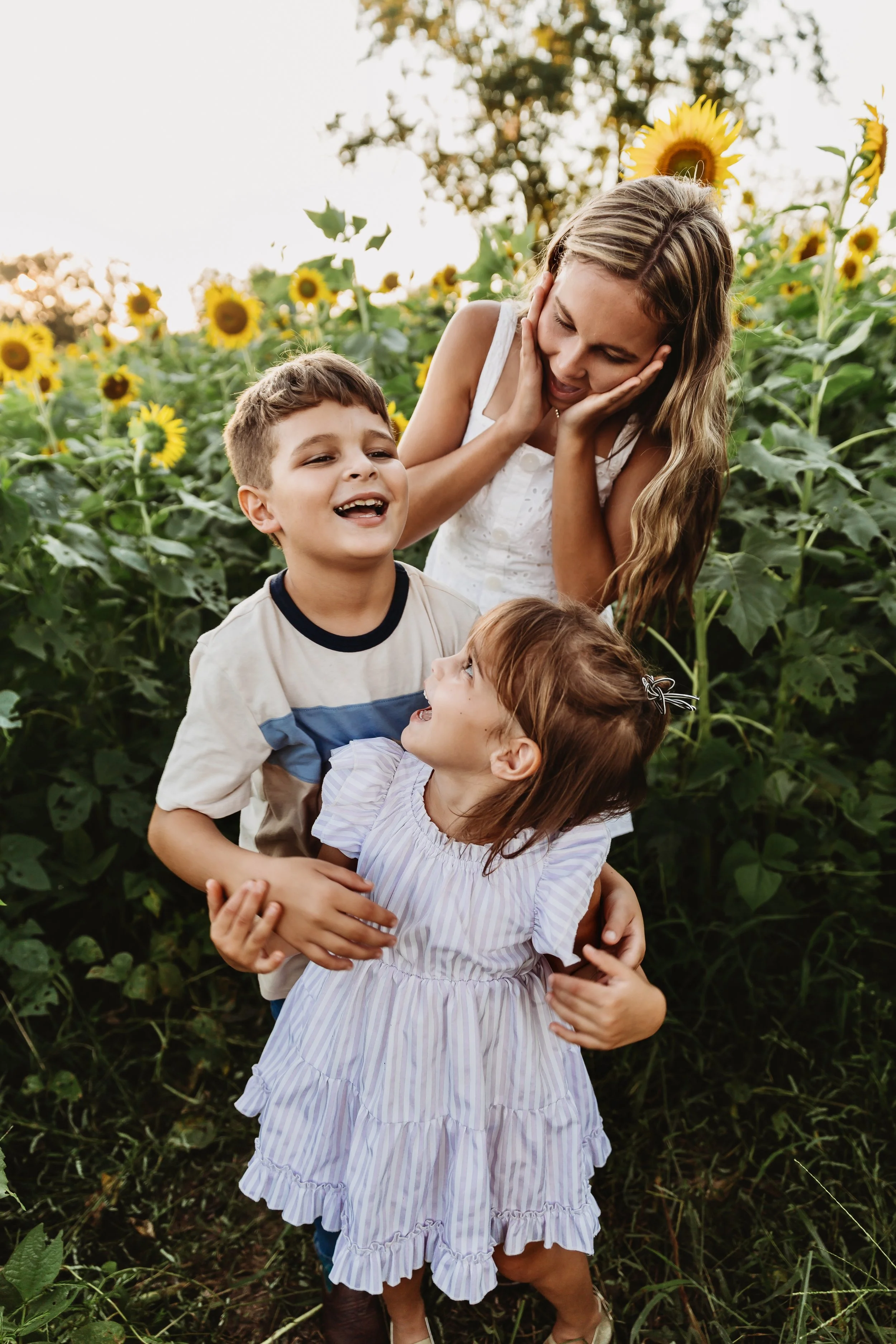 A woman and two children, a boy and a girl, standing in a sunflower field, enjoying each other's company. The woman is smiling and holding her face with her hands, while the children are laughing and looking at each other.