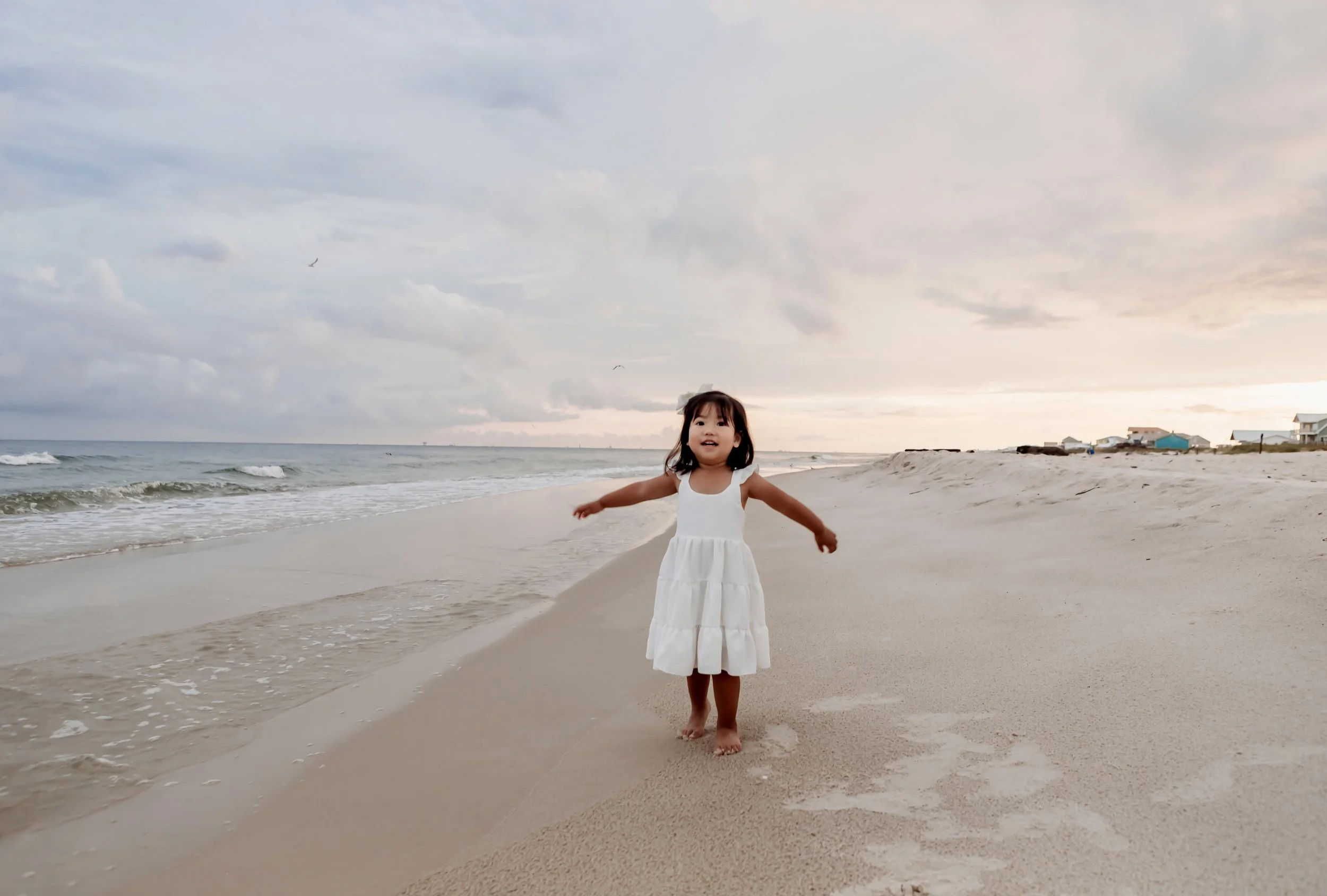 A young girl in a white dress standing barefoot on the sandy beach, smiling with arms outstretched, with the ocean and cloudy sky in the background during sunset.