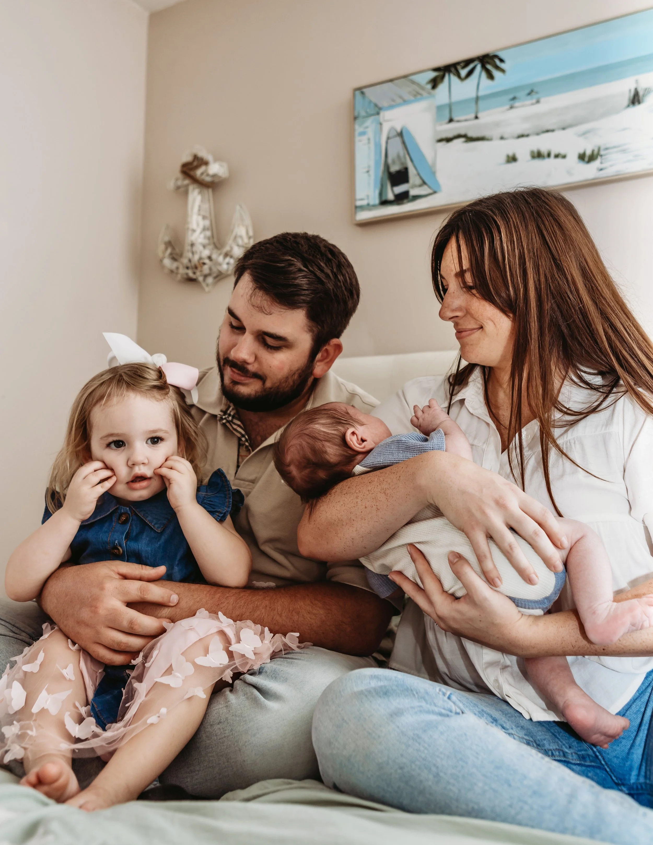 A family of four sitting on a bed in a bedroom, with two children and two adults. The mother is holding an infant, while the father is holding a young girl with a bow in her hair. They are all smiling and looking at each other.