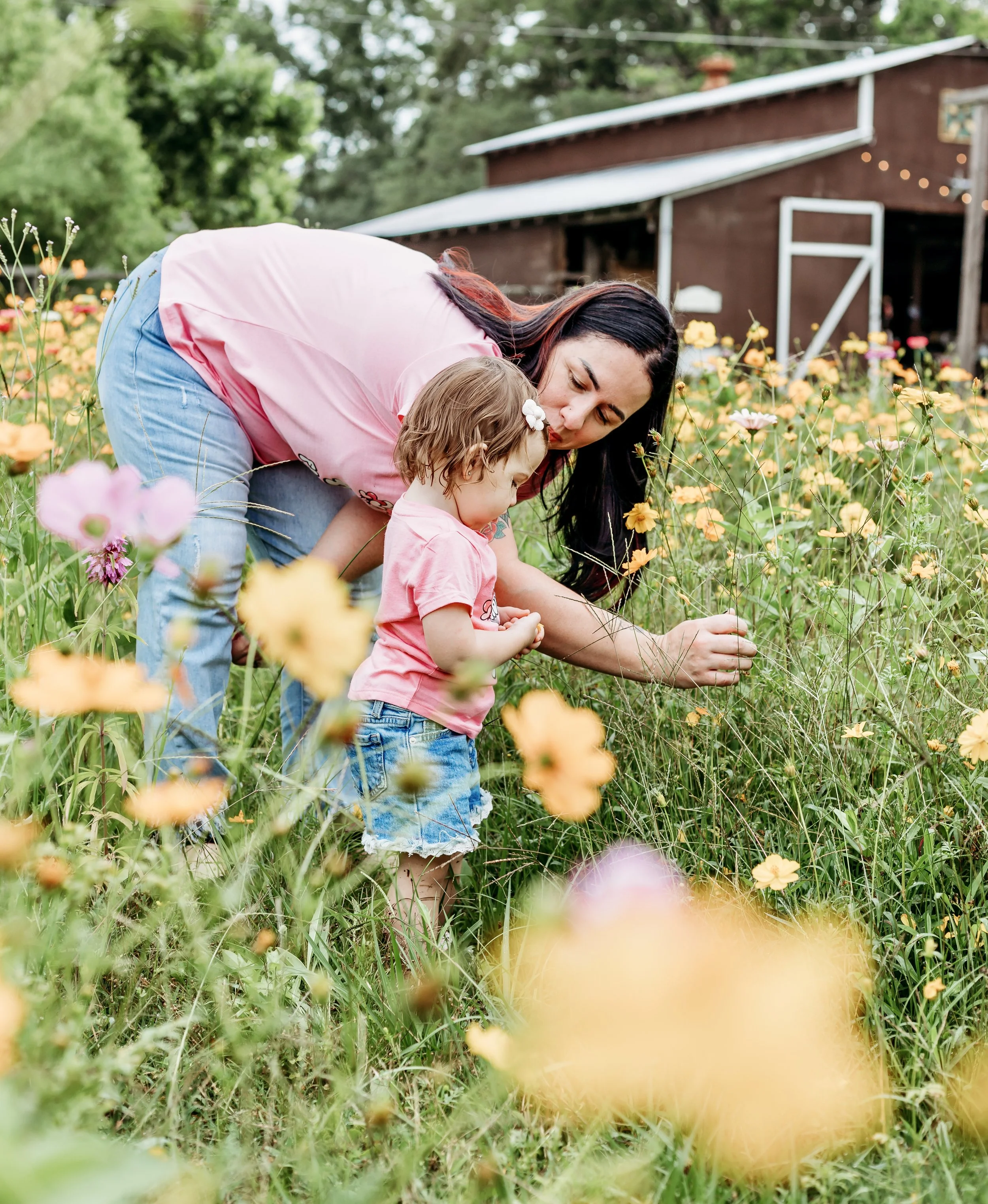 Woman and young girl picking flowers in a field of yellow and pink blooms outside a barn.
