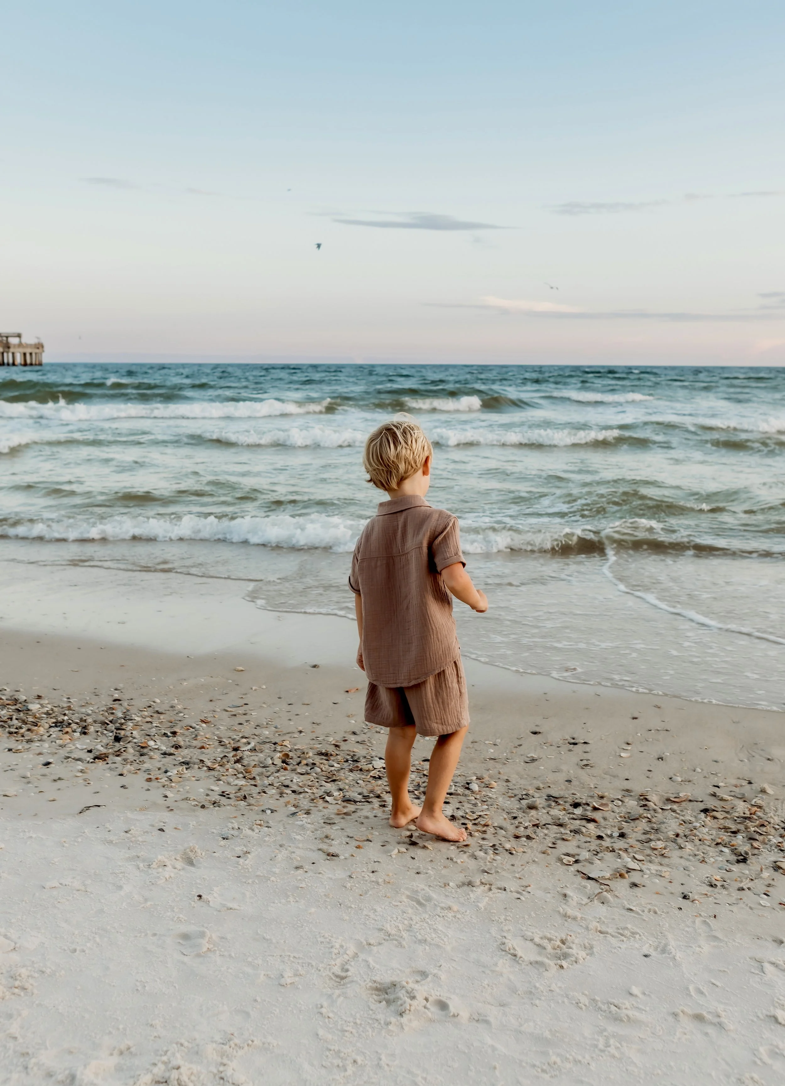A young boy standing barefoot on a sandy beach, looking out at the ocean waves during sunset or early evening.