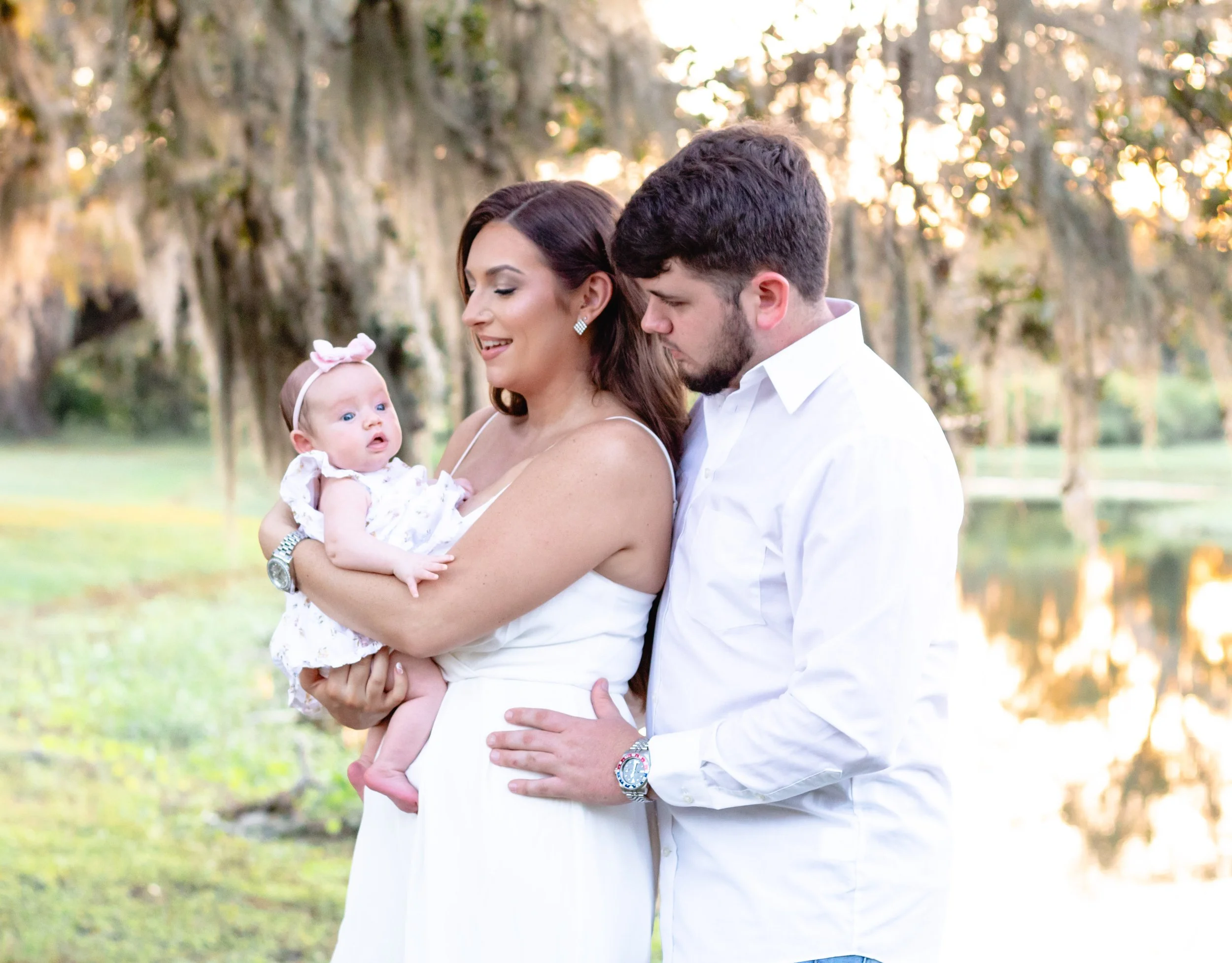 Family of three: woman holding a baby girl, man standing next to them, outdoors near a lake and trees, during sunset.