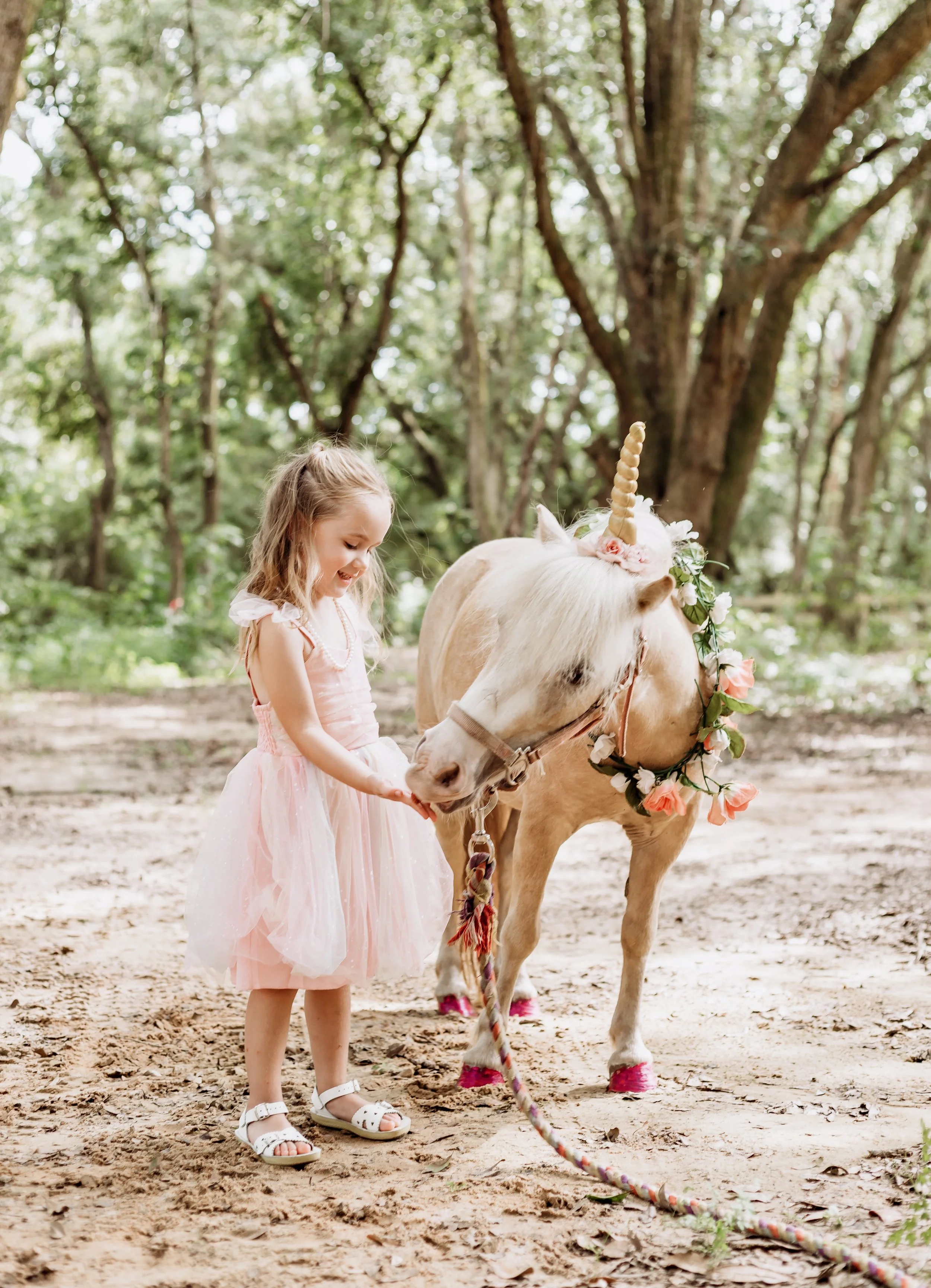 A young girl in a pink dress with a tulle skirt is petting a small unicorn. The unicorn has a golden horn, pink flowers around its neck, and pink shoes on its hooves. They are outdoors in a wooded area with green trees in the background.