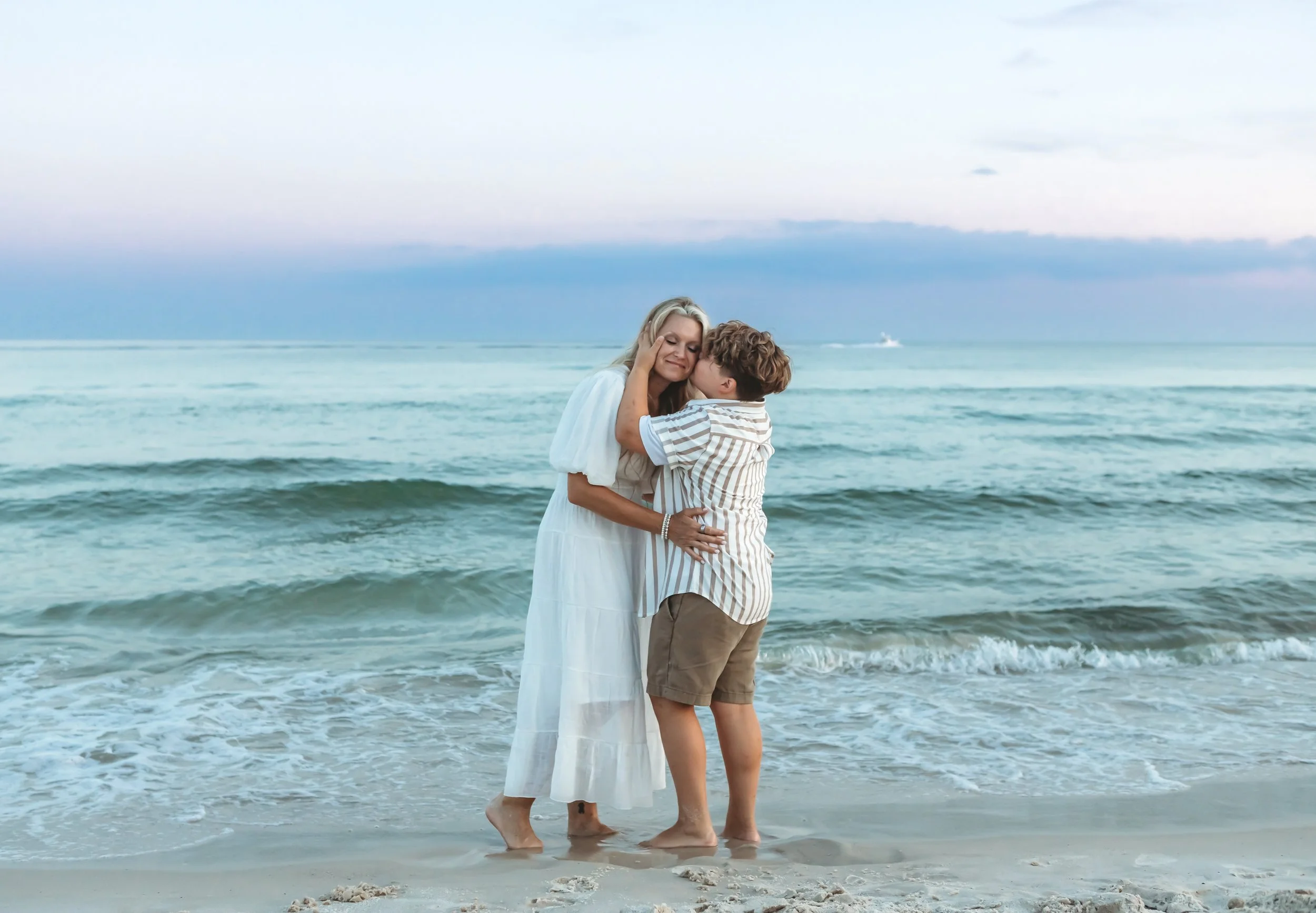 A woman and a young boy embrace and kiss on a beach at sunset or early evening with ocean waves in the background.