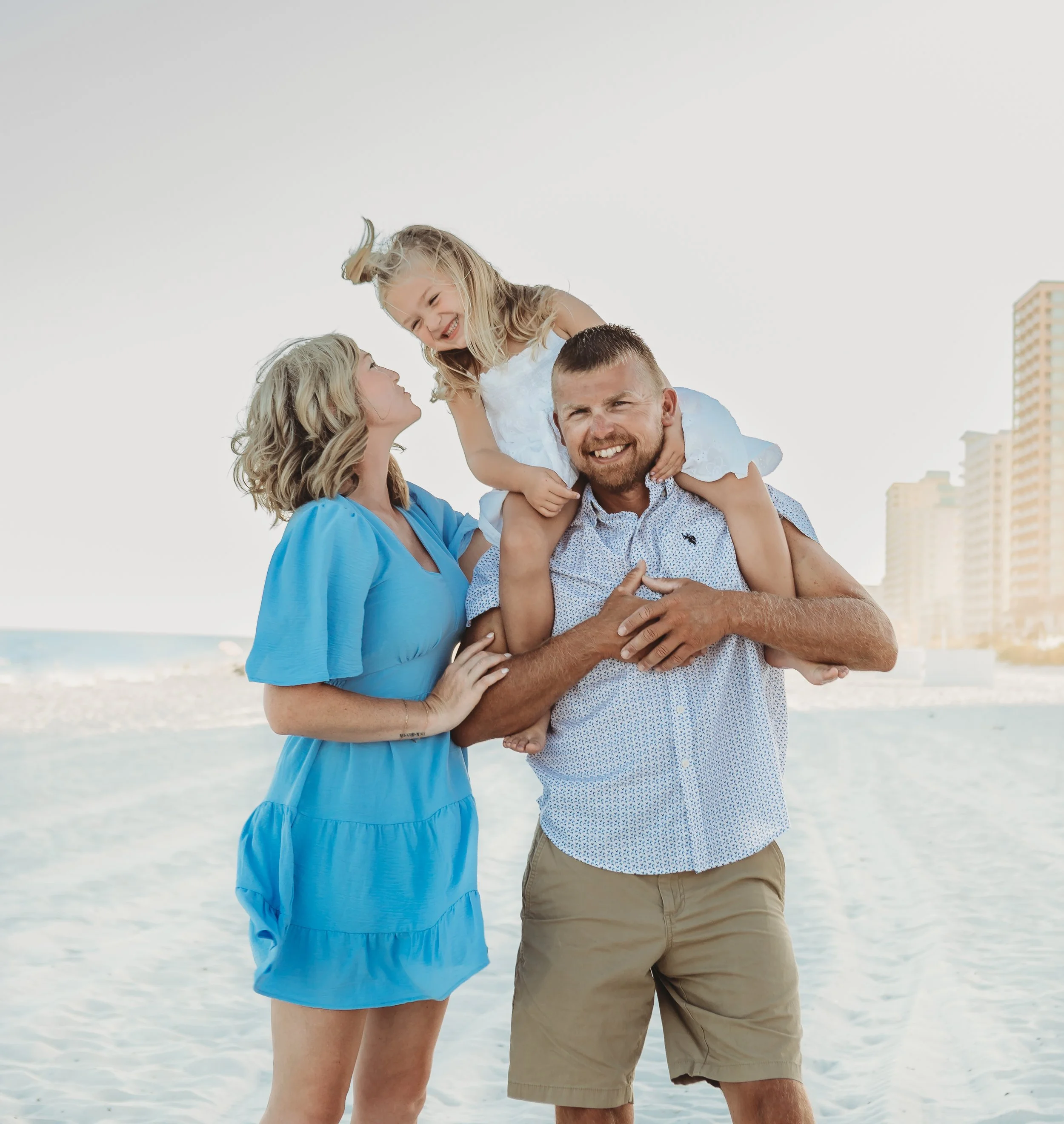 Family of four at the beach, with a man carrying a young girl on his shoulders, a woman beside them looking at her, and buildings in the background.