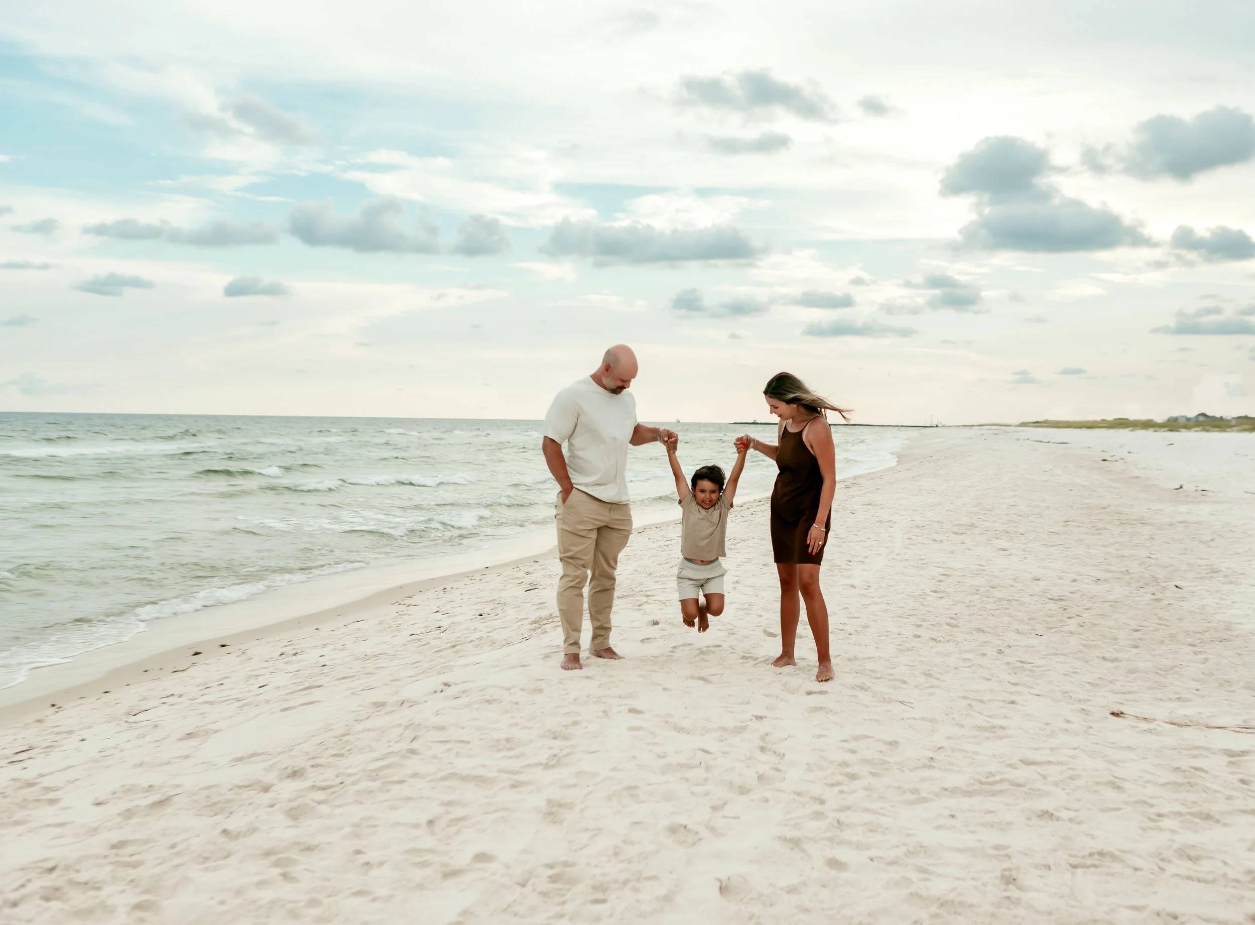 Family of three enjoying a day at the beach, with the father and mother holding the child's hands as she jumps in the sand, under a cloudy sky.