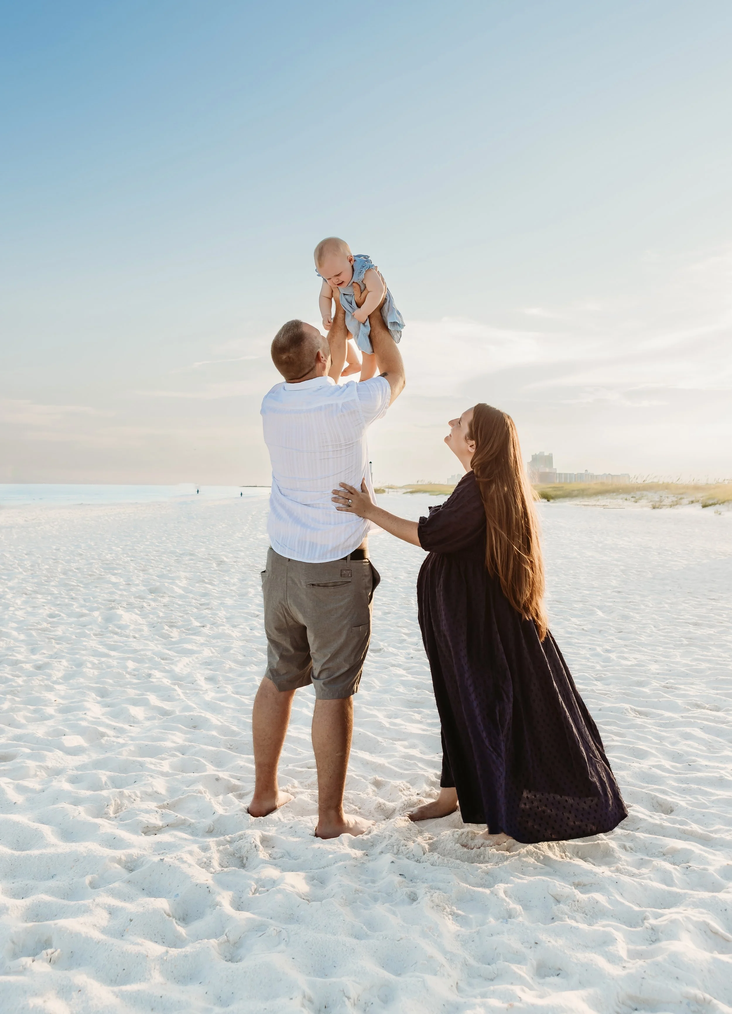 A family of three on the beach: a man holding a baby in the air, a woman kneeling and touching the man, with the baby smiling down at her. The sky is clear with a few clouds, and buildings are visible in the distance.