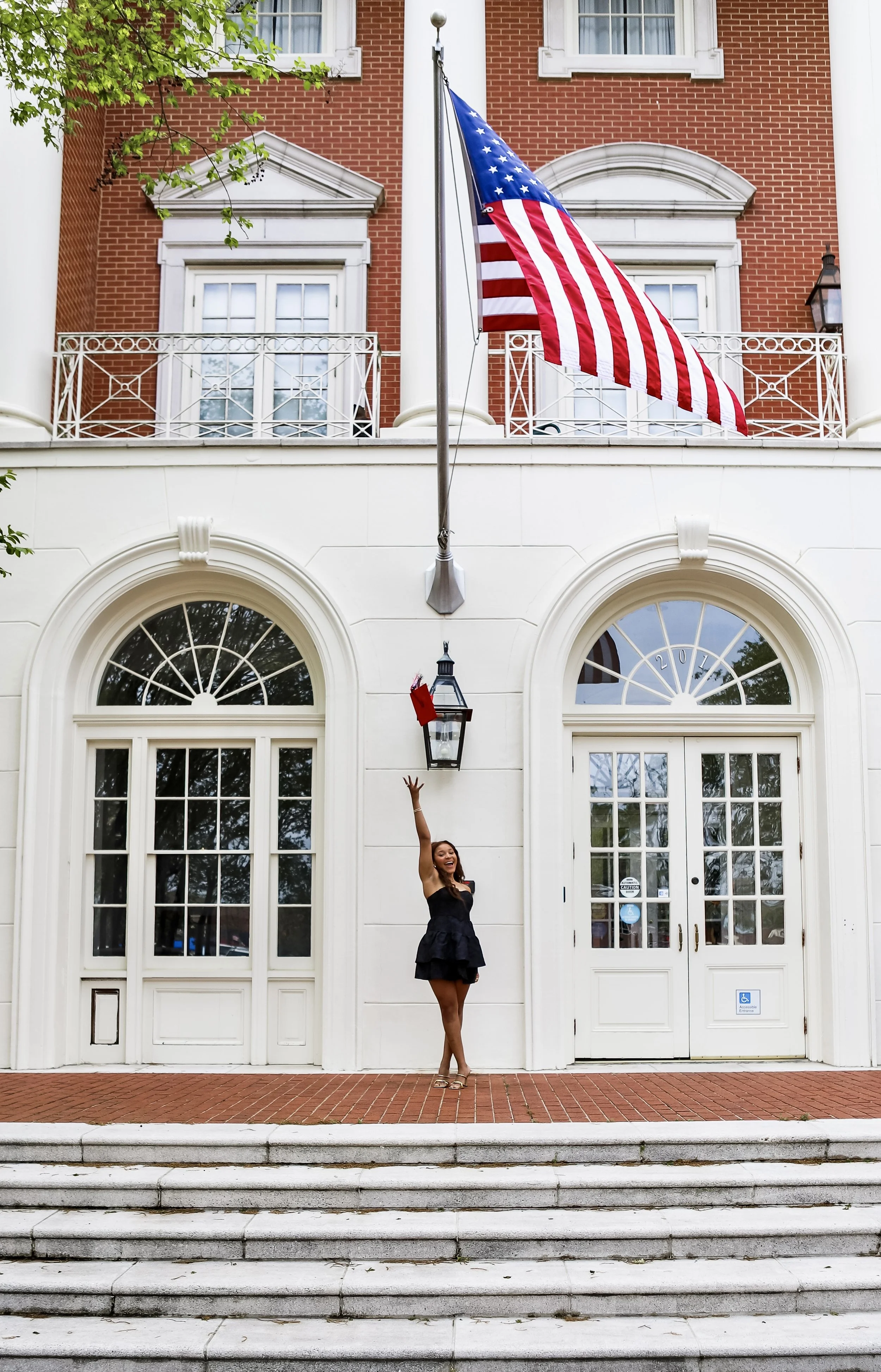 Woman in a black dress standing on steps in front of a white building, reaching up towards the American flag on a pole above her.