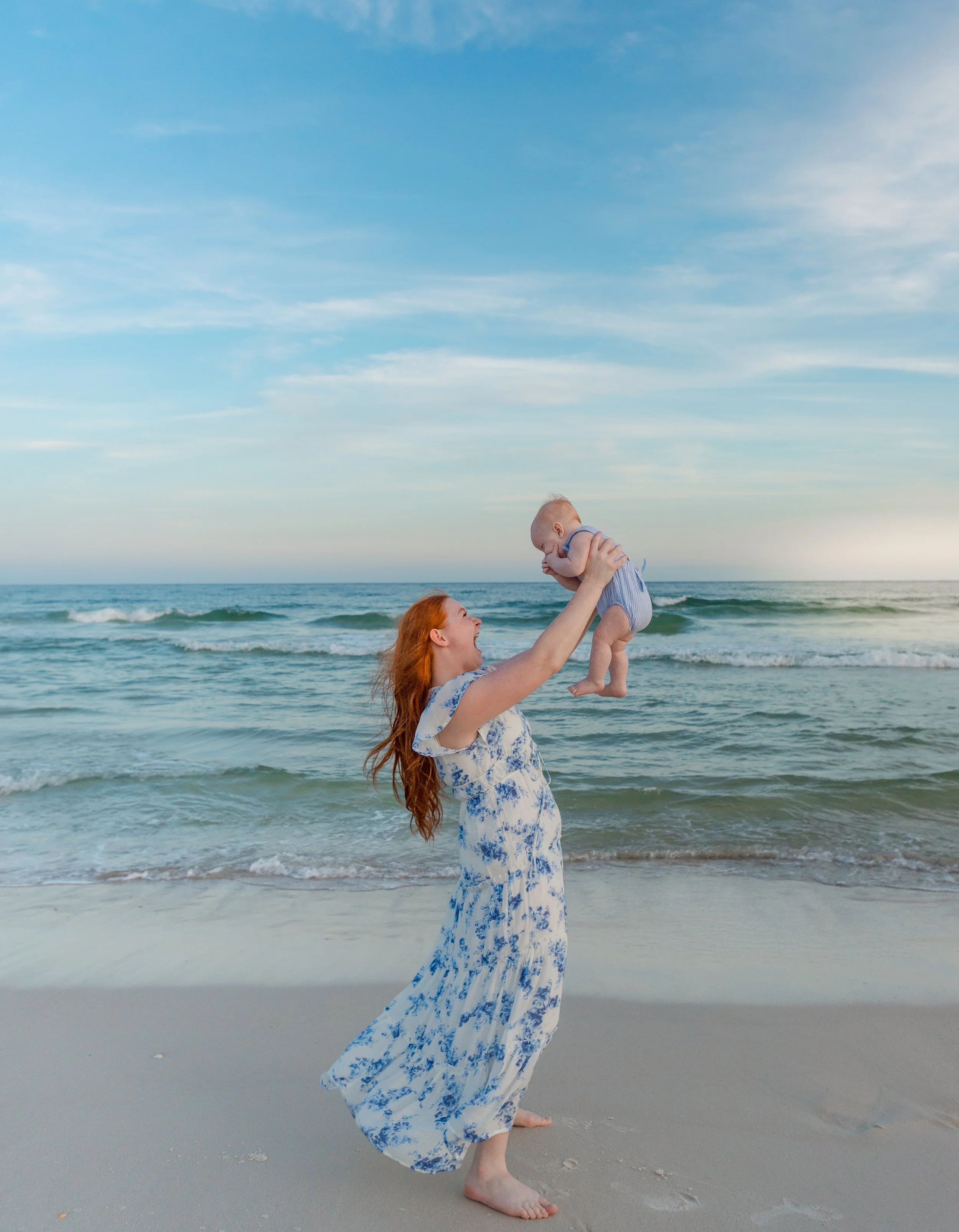 Young mom dressed in a blue flowy dress. Holding her baby boy up in the air. Enjoying the sunset in Orange beach Alabama 