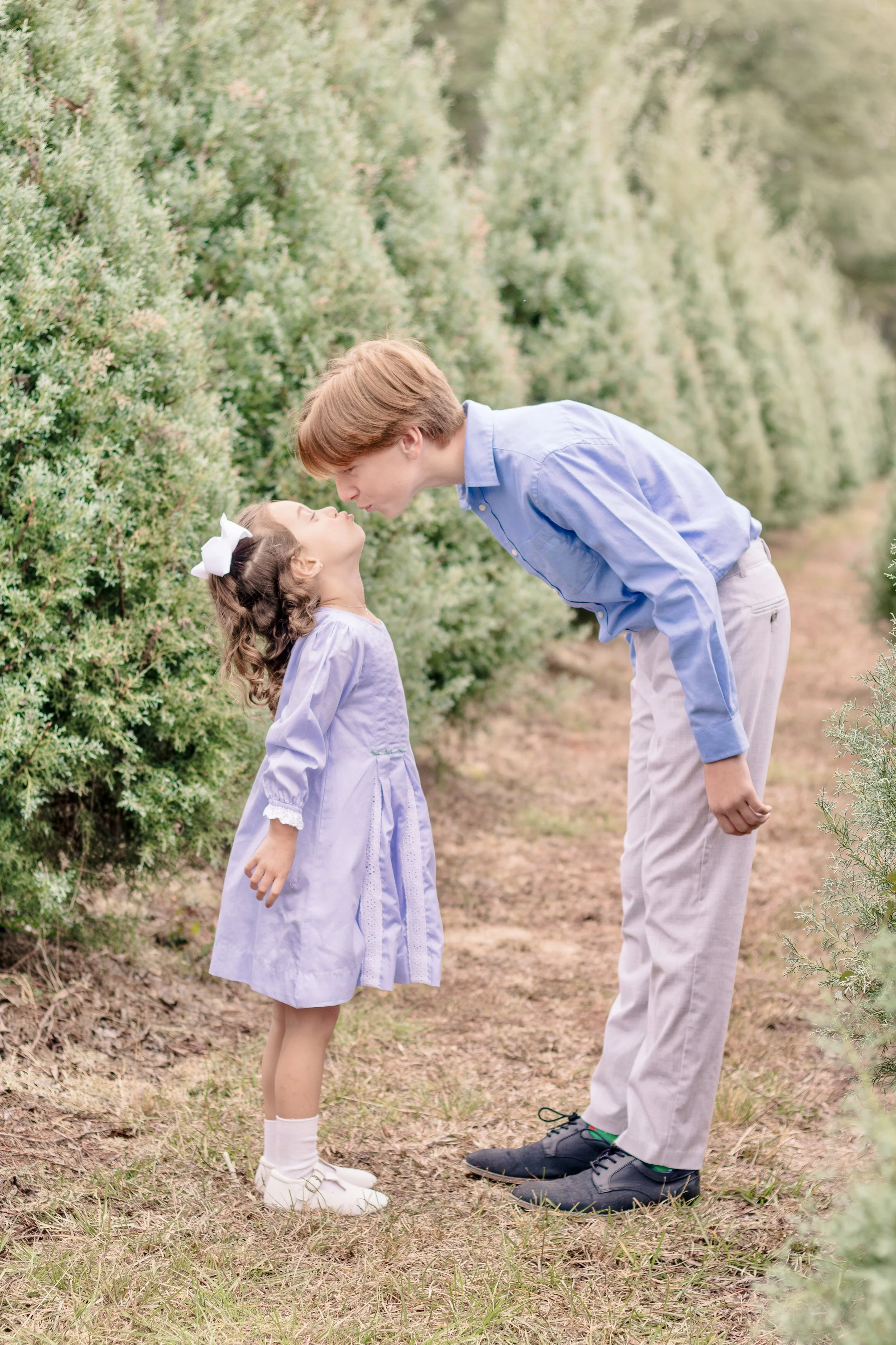 A young girl and an older boy stand close together on a dirt path, about to kiss amid green bushes.