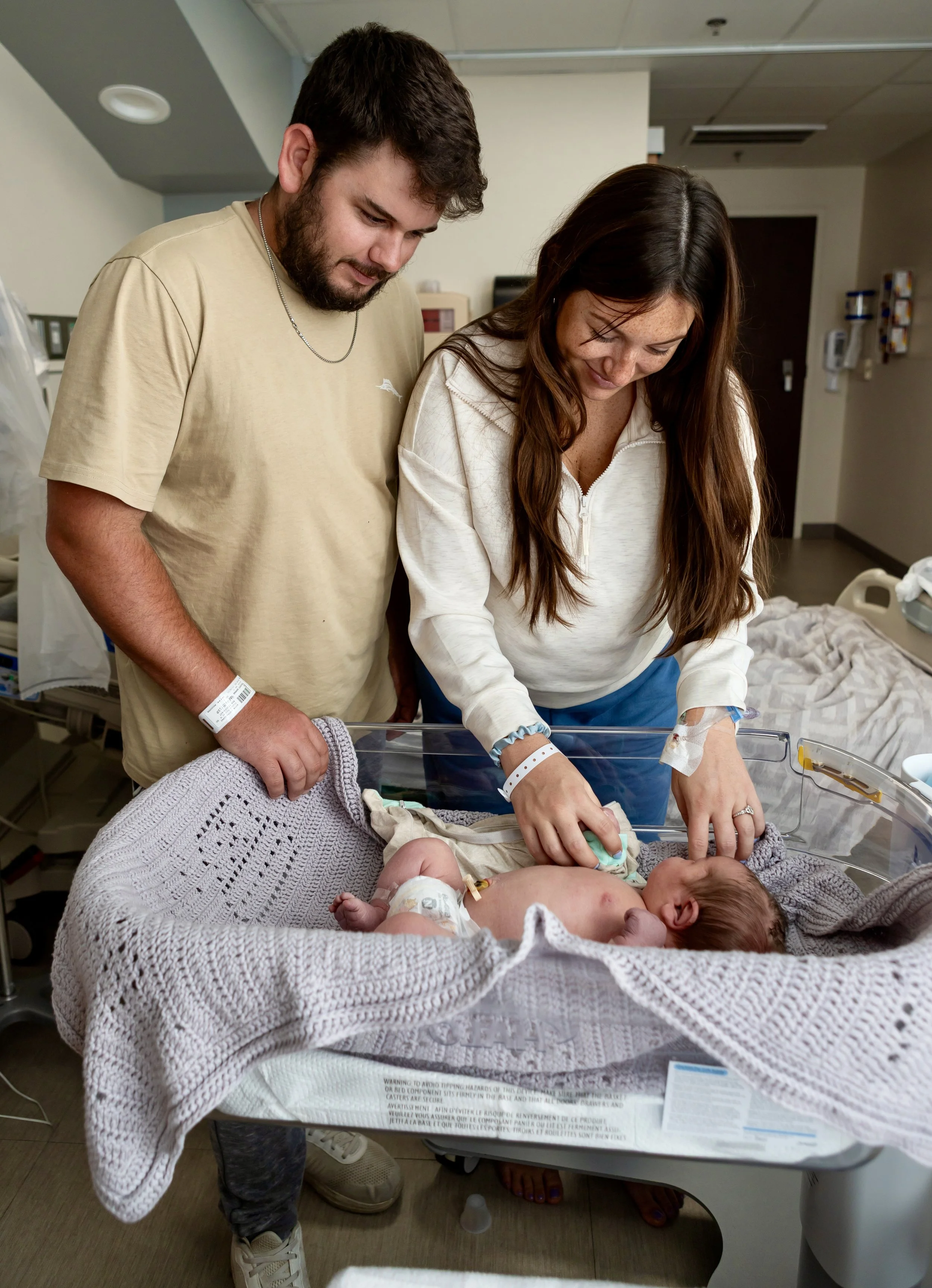 A new mother and father in a hospital room with their newborn baby in an incubator. The mother, wearing a white house shirt, is feeding the baby, while the father, in a beige t-shirt, stands beside her watching.