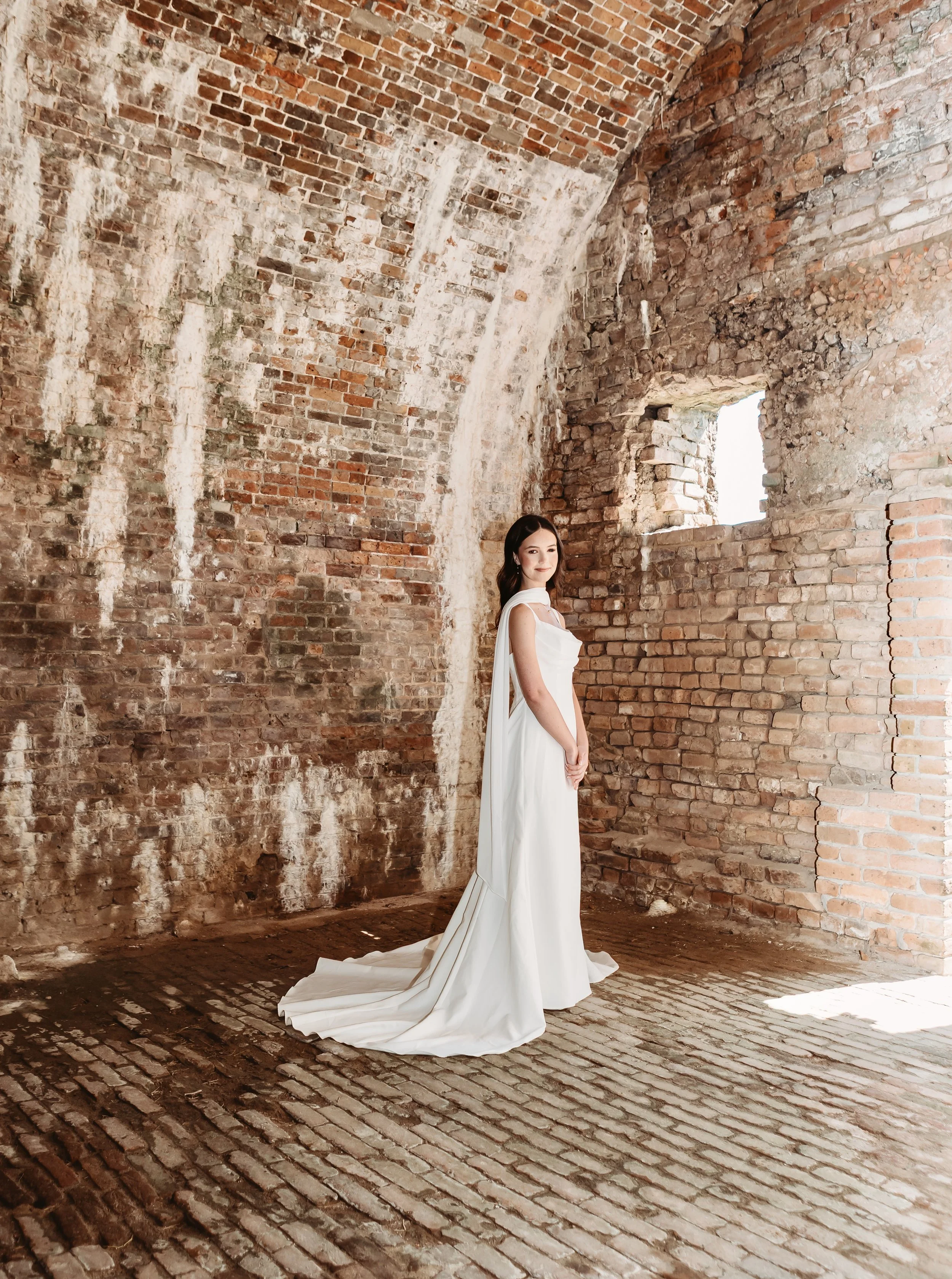 A woman in a long white dress standing in a rustic brick room with a small window, with textured and weathered brick walls and floor.