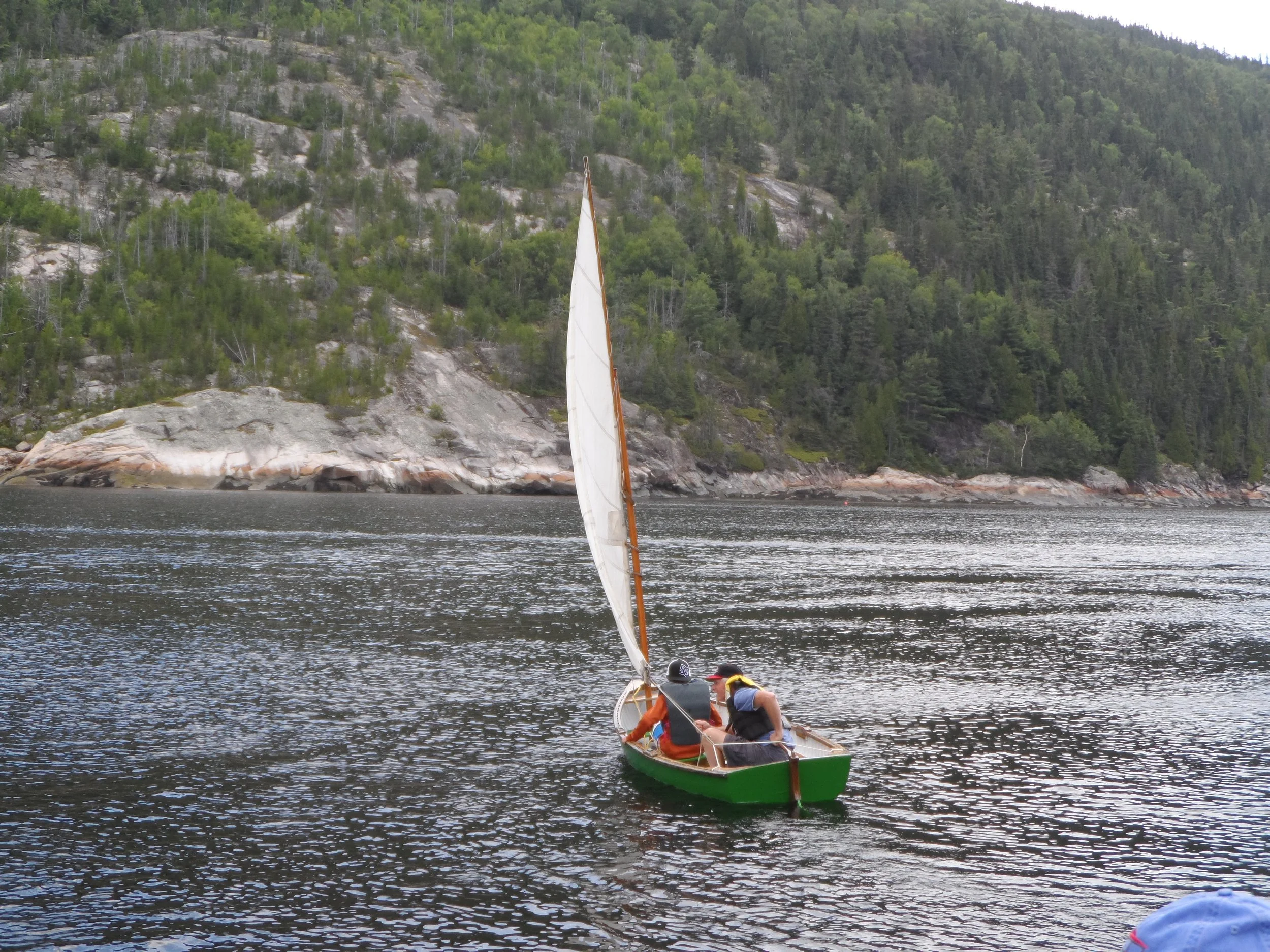 Two people in a small green sailing dinghy on the Saguenay River near a forested shoreline with rocks and trees.