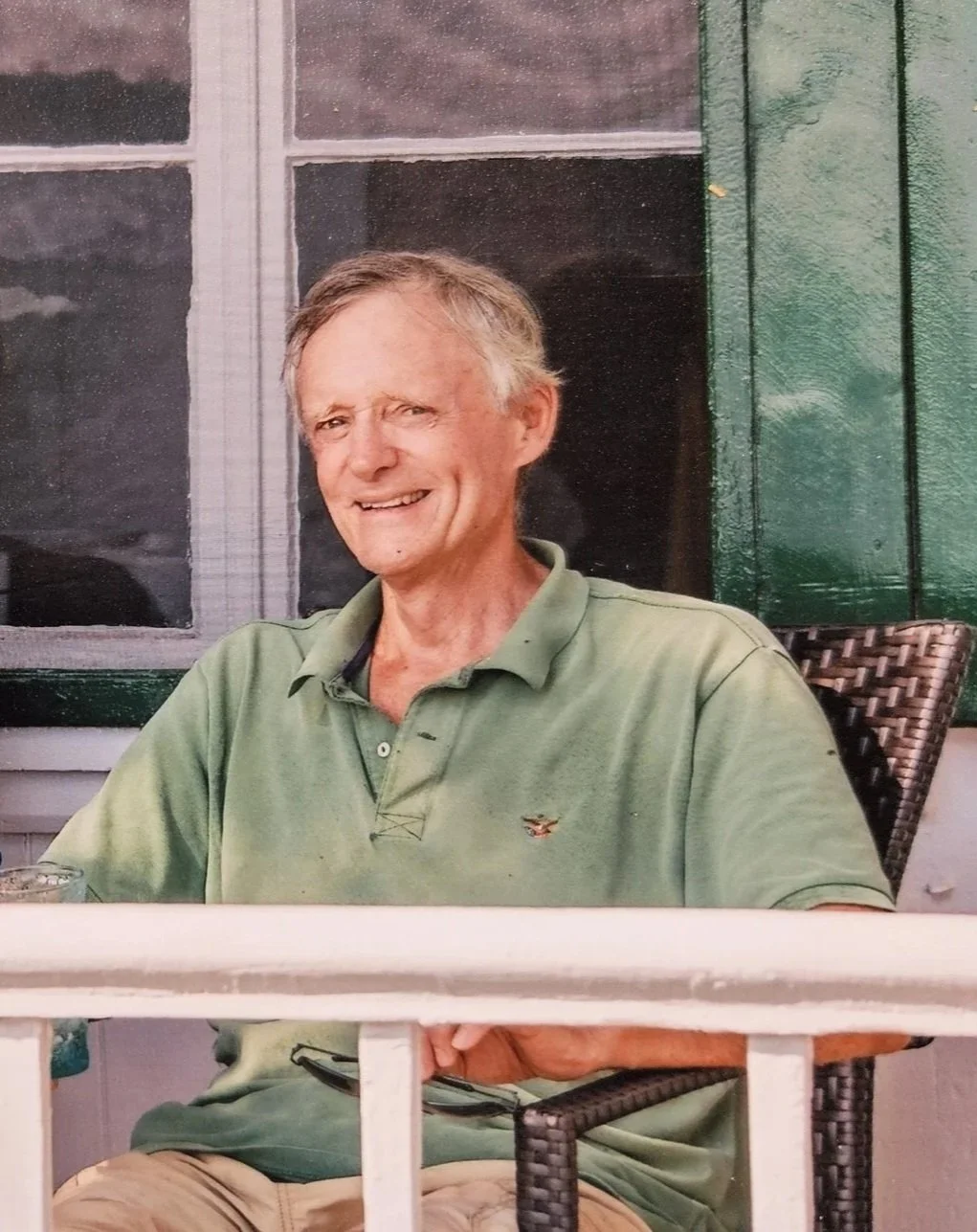 Alan Morewood Evans, wearing a green polo shirt, sitting on a porch with a green-shuttered window in the background.