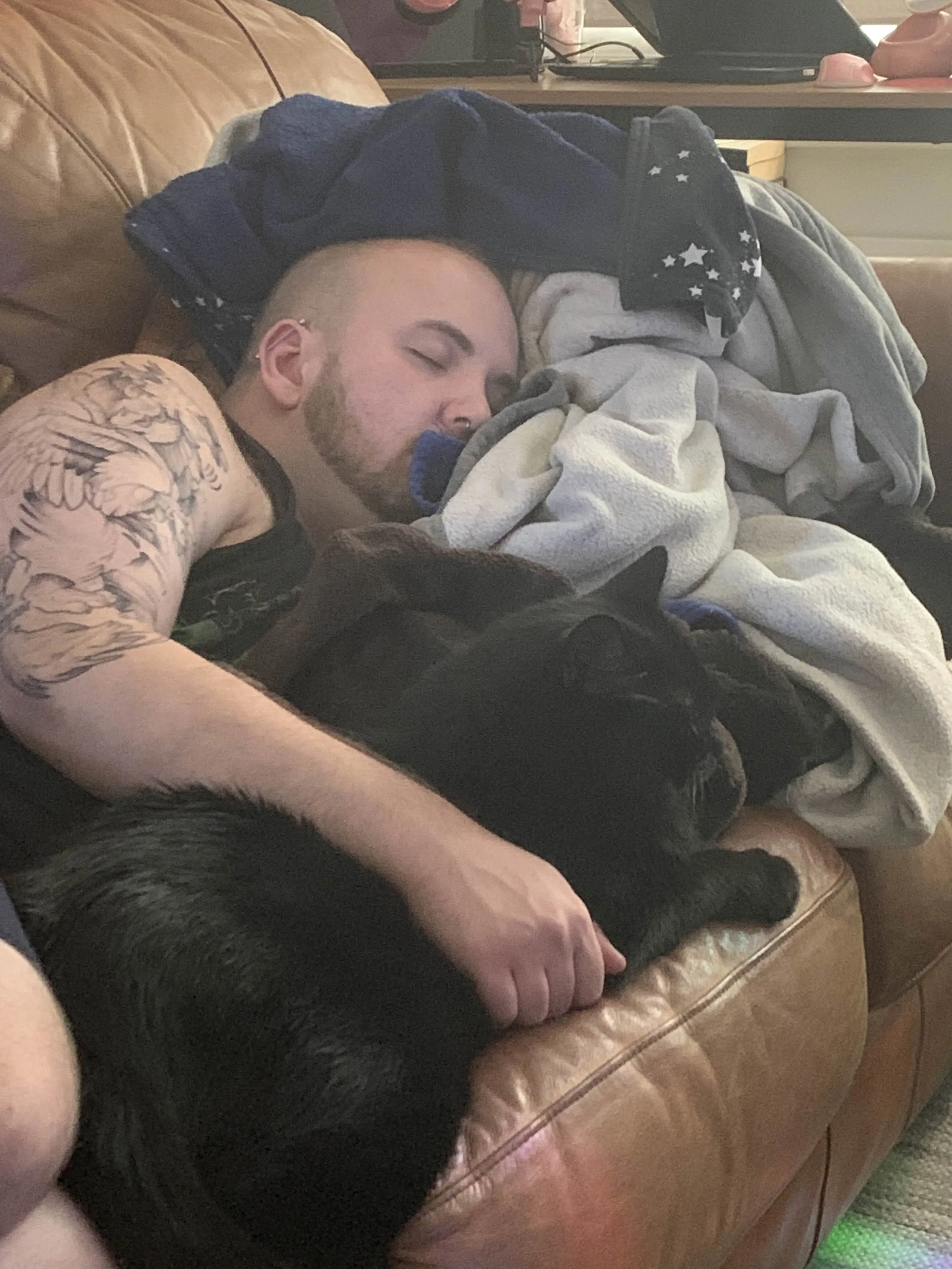 Lowell, a white nonbinary person with a shaved head and short beard. They are asleep on a couch holding an enormous black cat as if he's the little spoon.