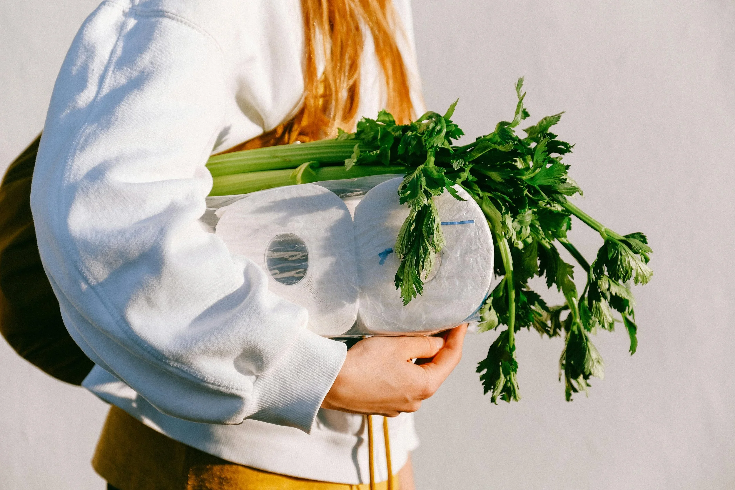 Person holding a plastic bag filled with green vegetables, including celery and leafy greens, against a plain background.