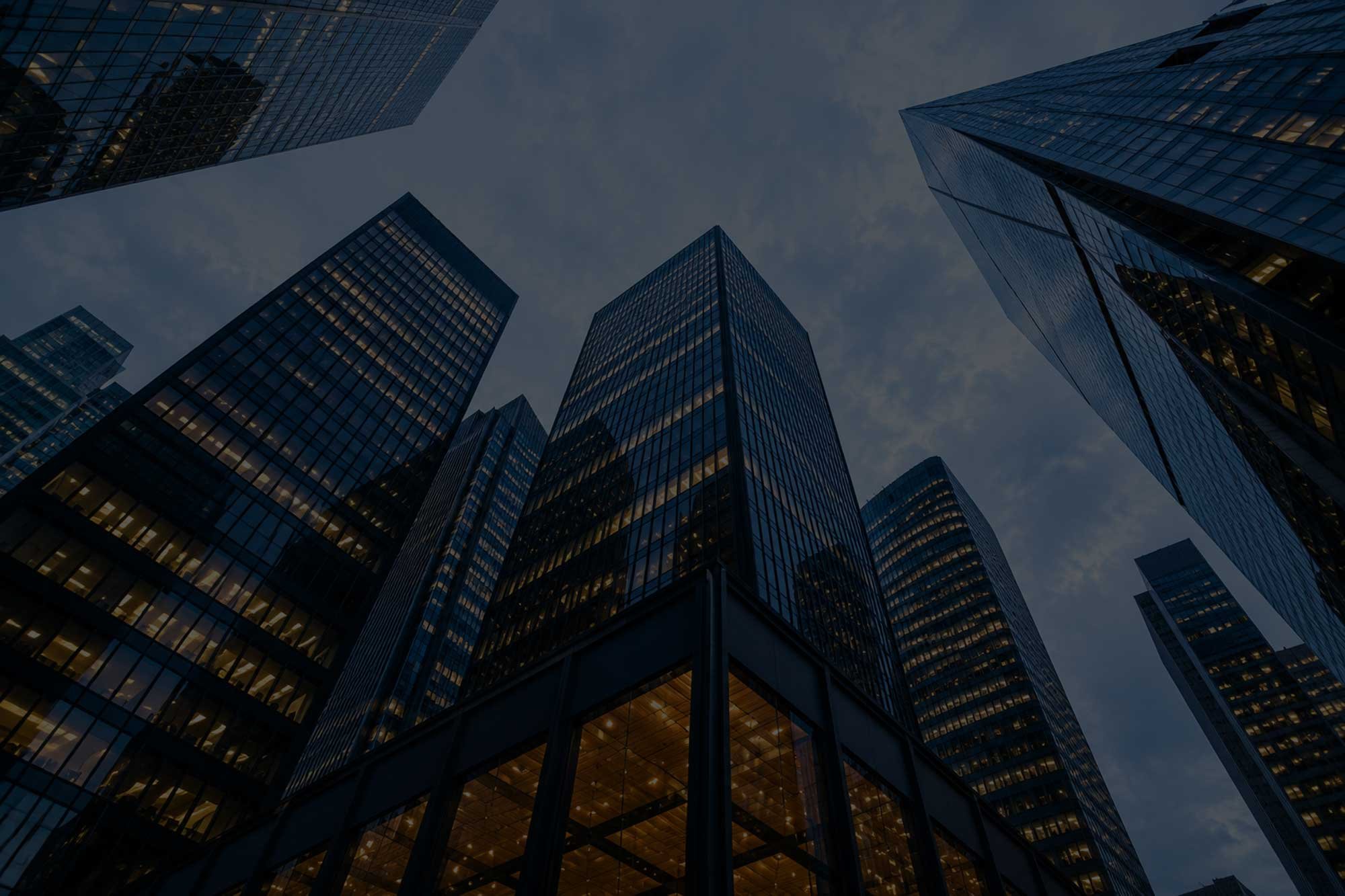 Looking up at tall glass skyscrapers in a city during dusk, with lights inside the buildings beginning to turn on, reflecting a darkening sky.