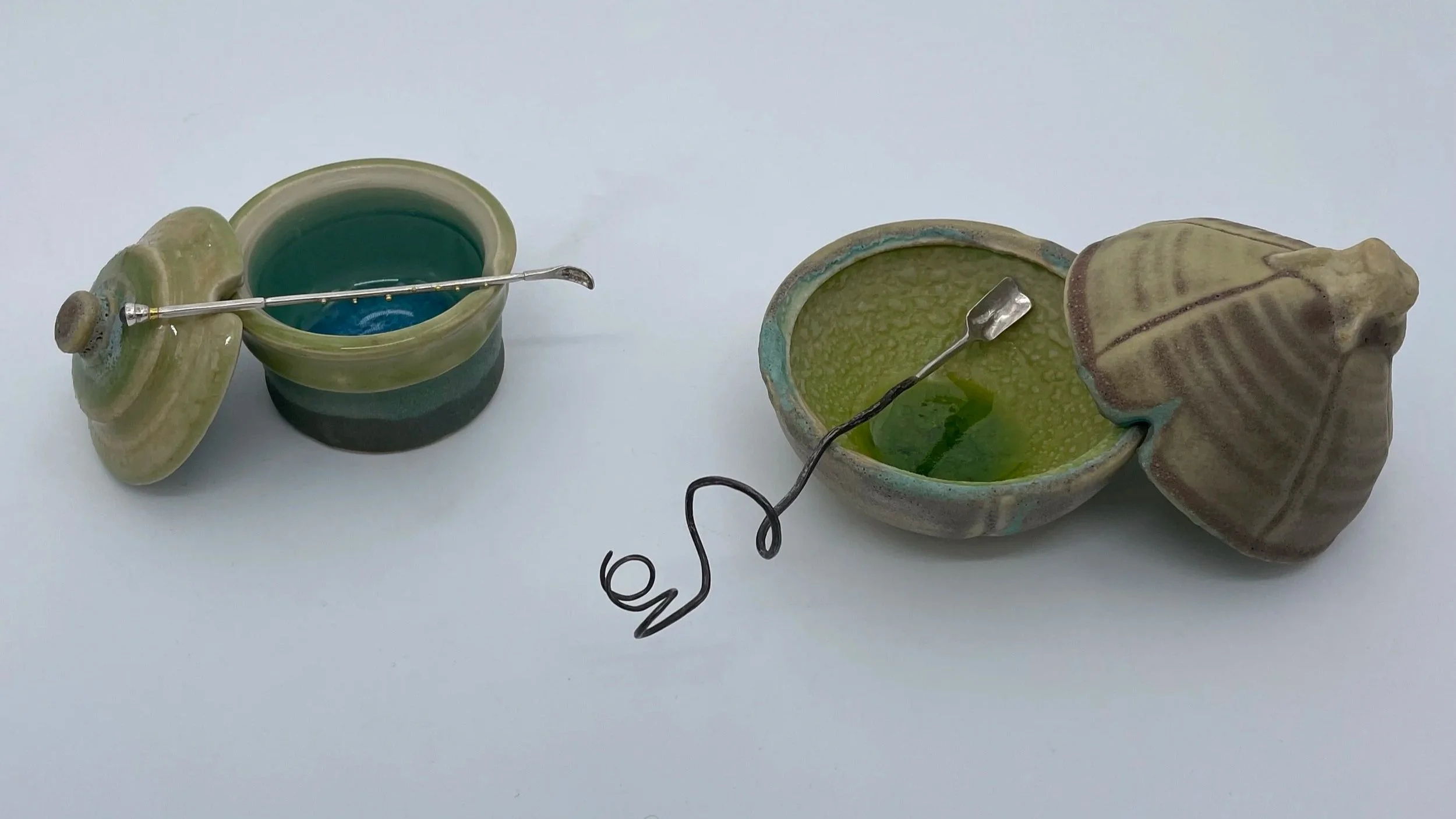 Two ceramic containers with lids and small metal utensils inside, one containing a green glaze and the other with a blue glaze, placed on a white surface.