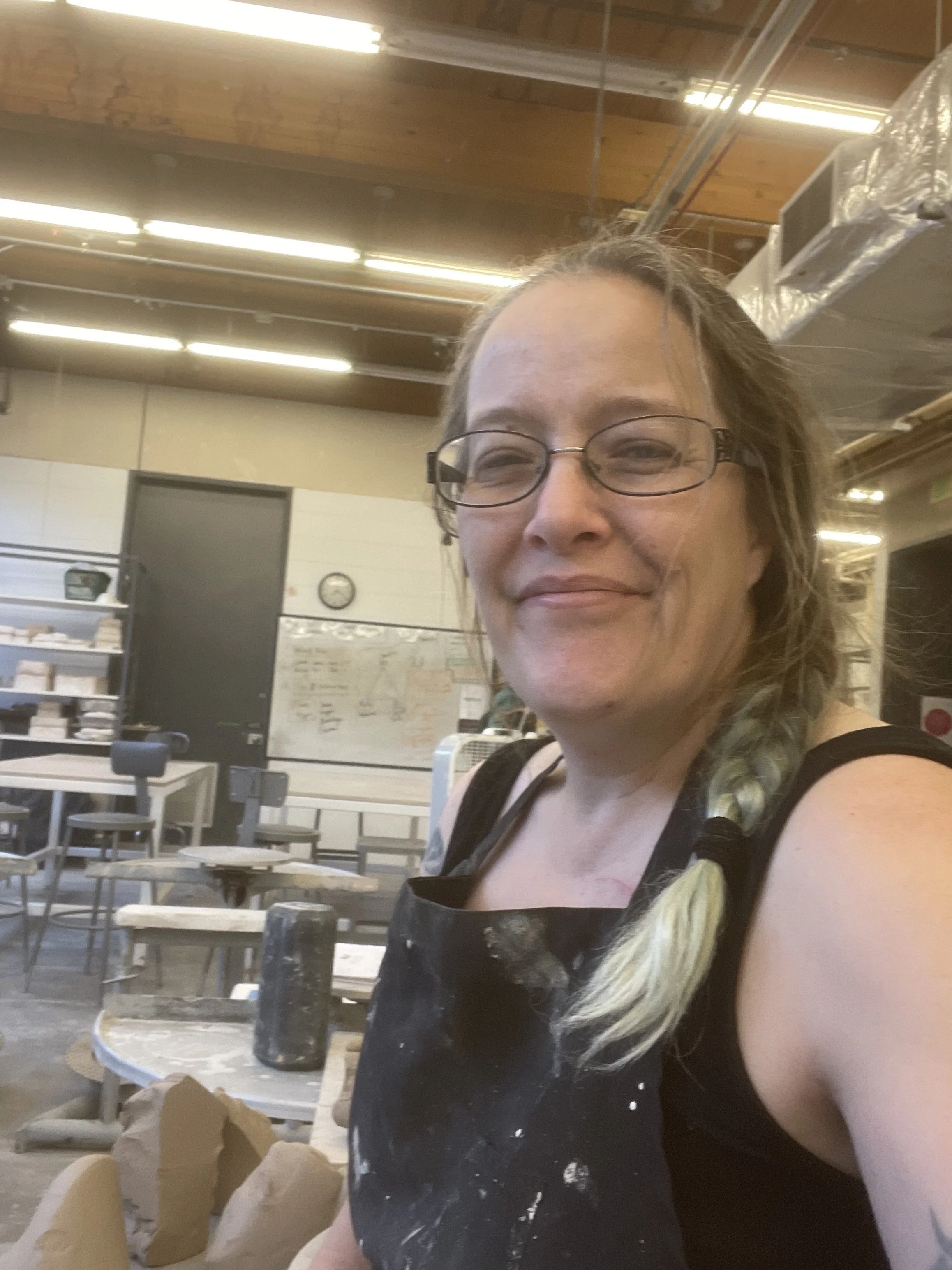 The artist, a woman with glasses, light brown hair with gray streaks, and a smile, working in a pottery studio with pottery wheels around her.