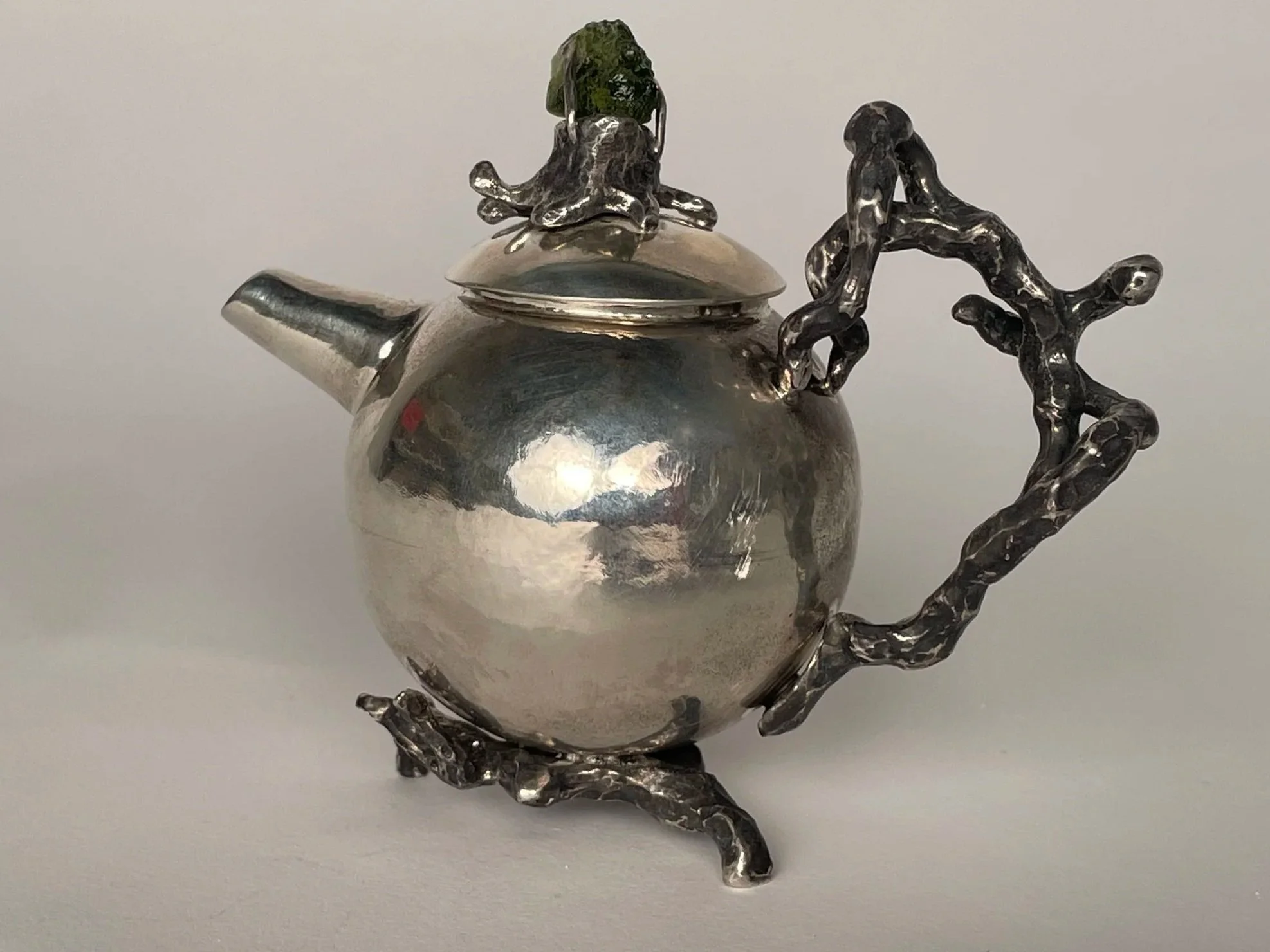 A sterling silver teapot with an ornate, twisted metal handle and spout. The teapot has a sculptural decorative element on the lid with a small green stone.