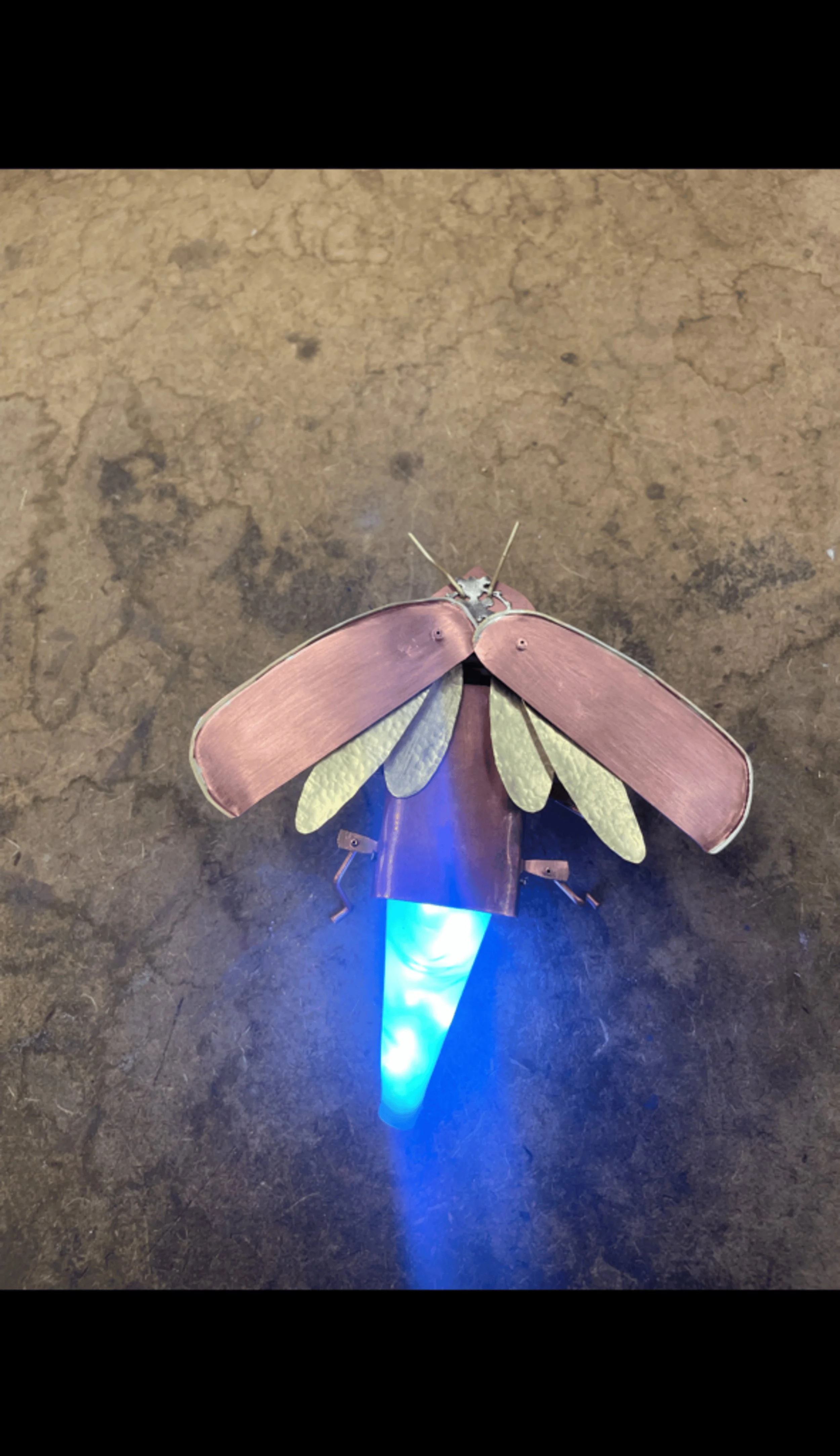 A decorative metal lightning bug with copper and brass wings mounted on an illuminated tail.