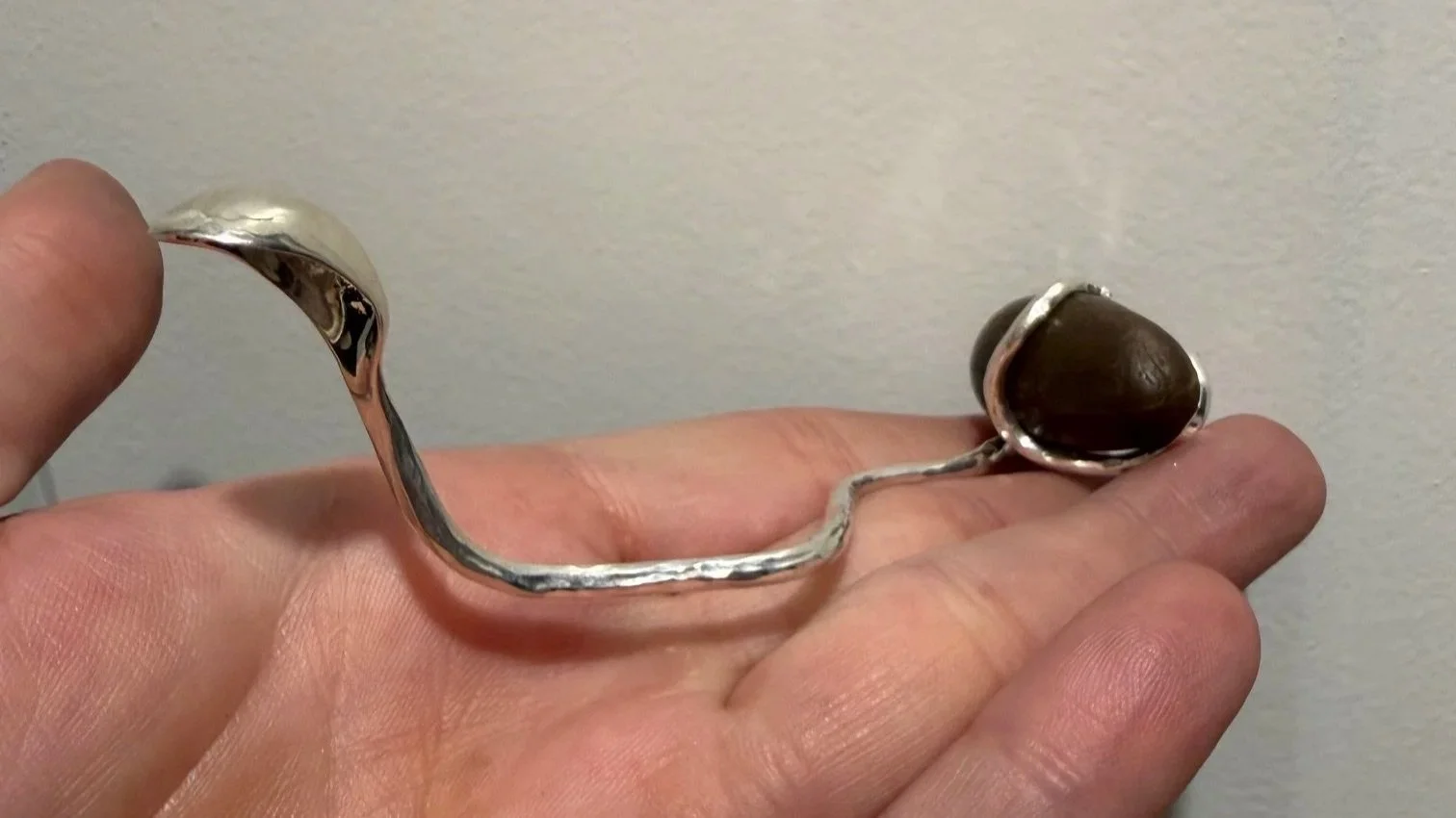 A person holding a silver snake-esque tiny spoon with a dark brown stone and twisted design.