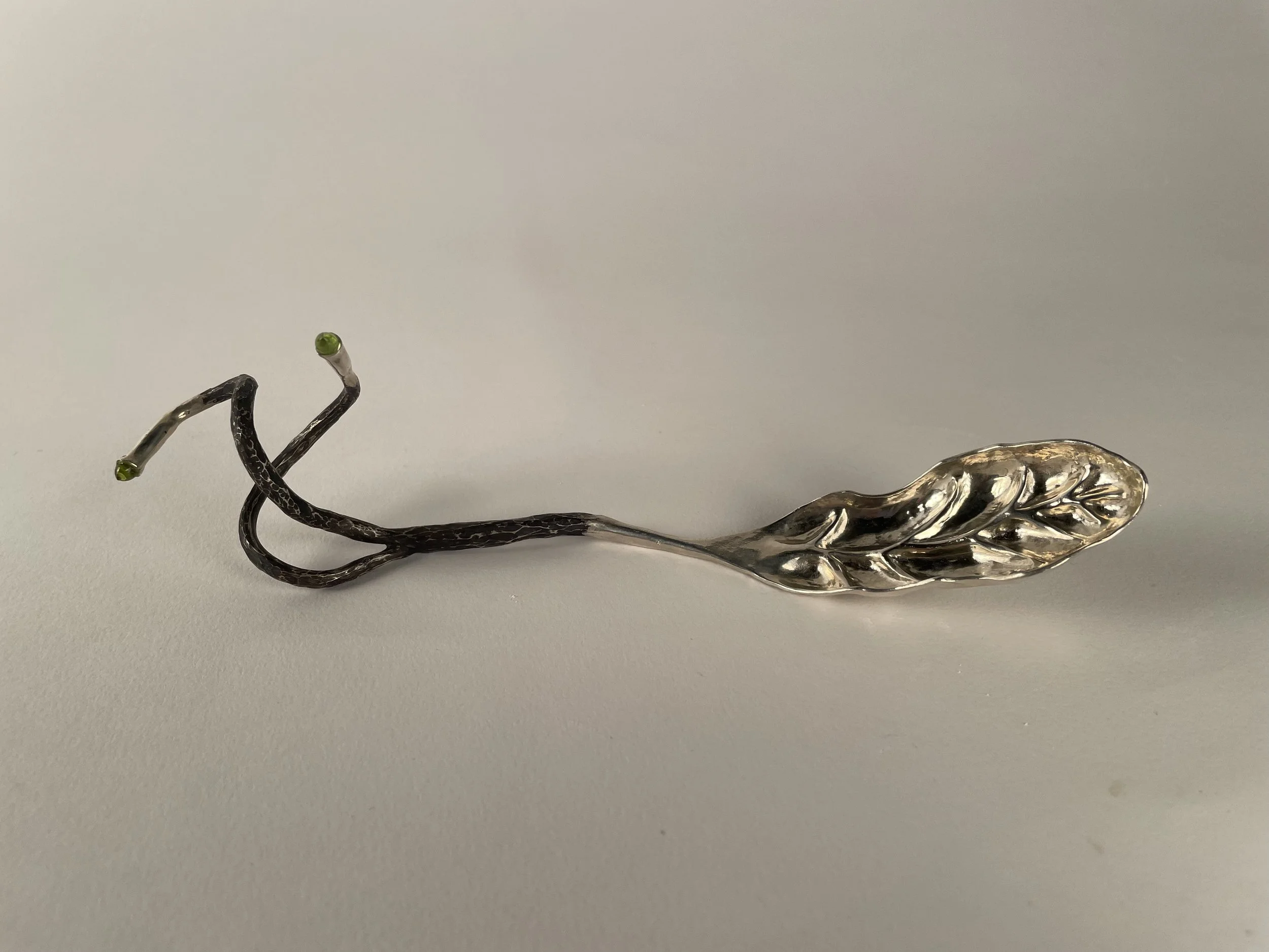 A decorative, hand forged, chased and repoussé sterling silver spoon, with small peridots at the tips of its twigs, placed on a plain white surface.