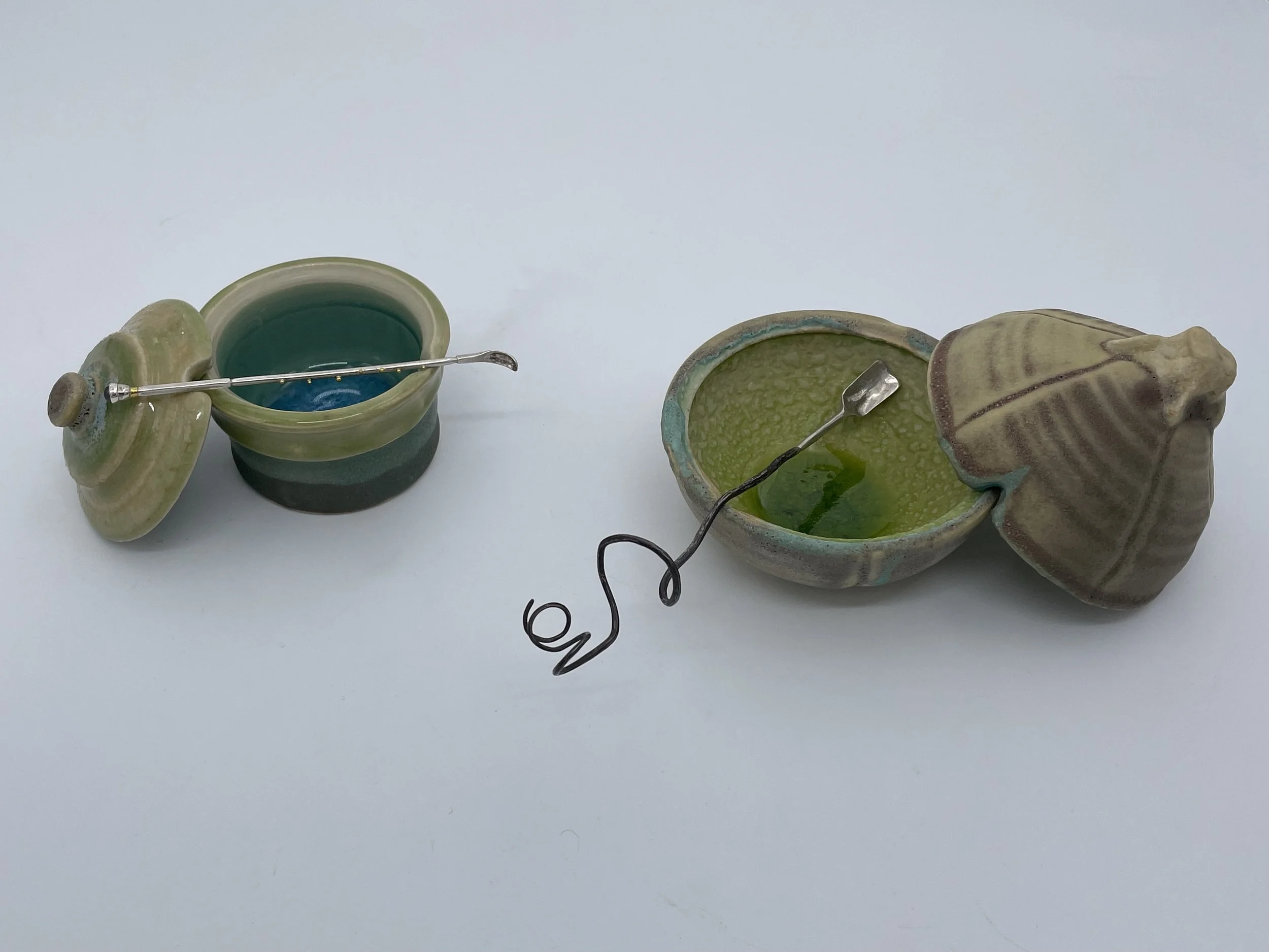 Two ceramic salt pots, with accompanying and complimenting hand forged spoons, on a white surface.