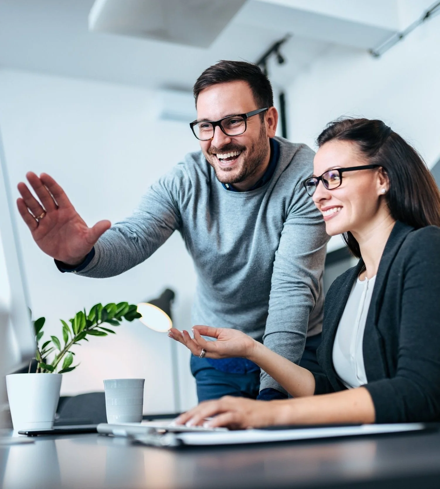 Two colleagues, a man and a woman, smiling and discussing work in an office. The man is leaning over and waving, while the woman is sitting at a desk with a potted plant nearby.