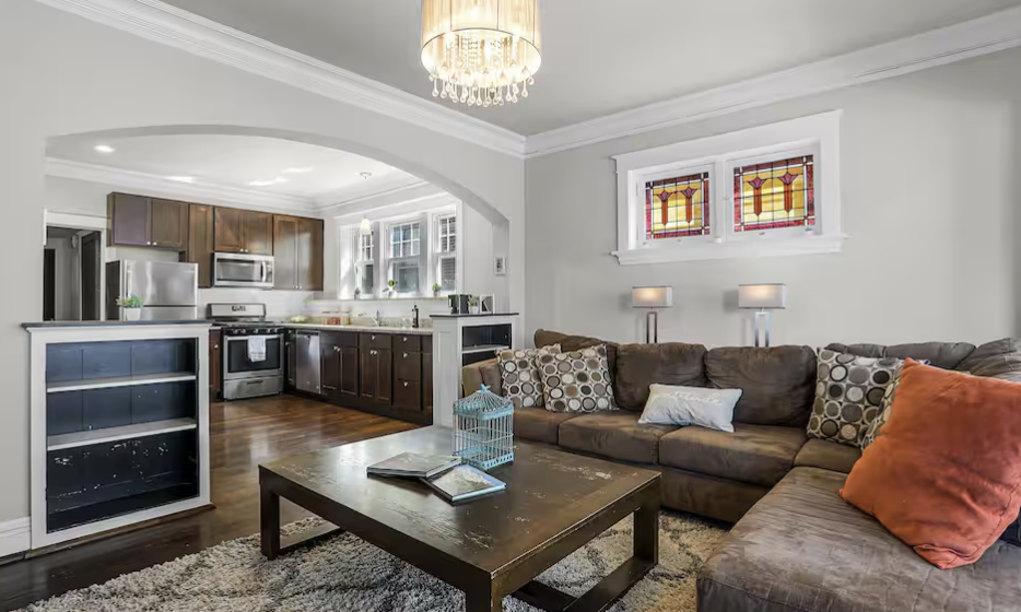 Living room with brown sectional sofa, orange and patterned throw pillows, wooden coffee table, and two lamps on the wall. Adjacent kitchen with dark wood cabinets, stainless steel appliances, and a windowed wall.