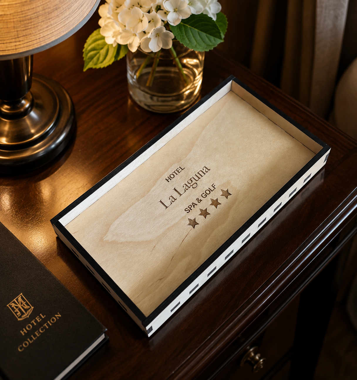 A wooden desk with a black and white framed hotel information card reading 'Hotel La Laguna Spa & Golf' with five stars, a black guestbook labeled 'Hotel Collection,' a brass lamp, and a glass vase with white flowers.