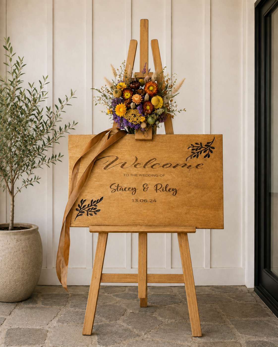 Wedding welcome sign on an easel with a floral arrangement and a potted plant beside it.