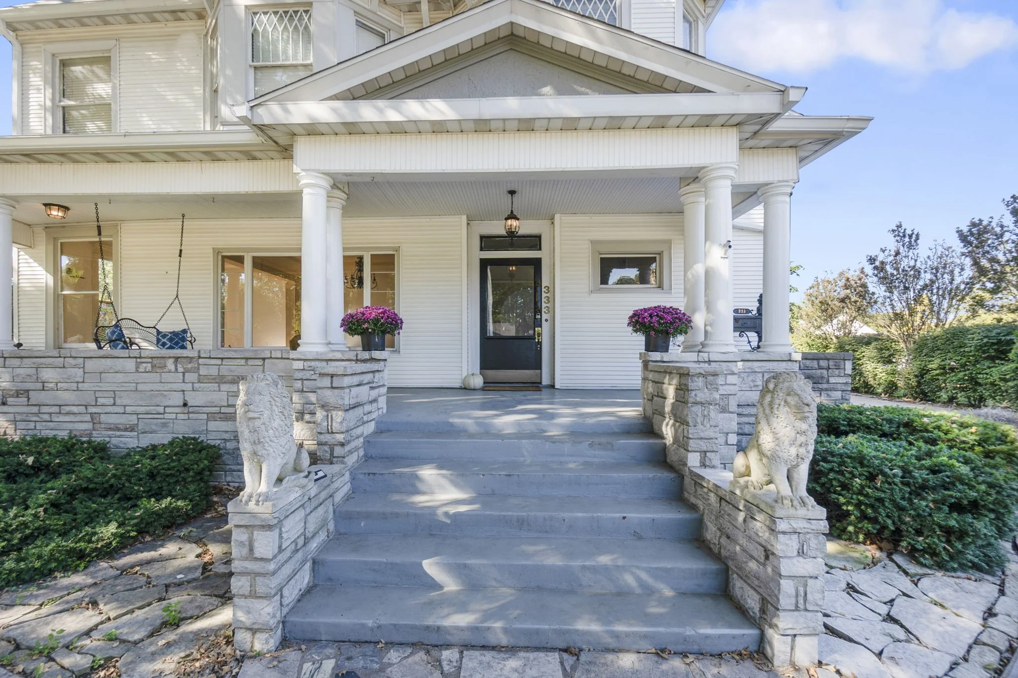 Front porch of a white house with columns, potted pink flowers, stone lions, and steps leading up to the black front door.