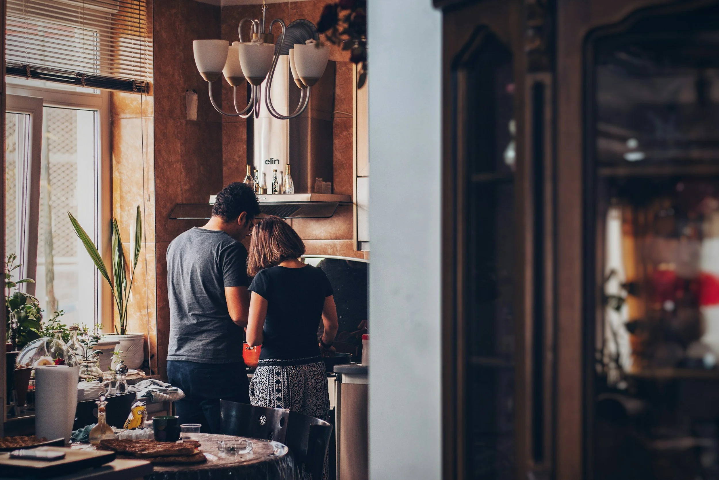 A man and woman cooking together in a cozy kitchen. The man is wearing a gray t-shirt, and the woman is wearing a black top and patterned pants. The kitchen has wooden cabinets, hanging wine glasses, and a window with blinds allowing natural light in.