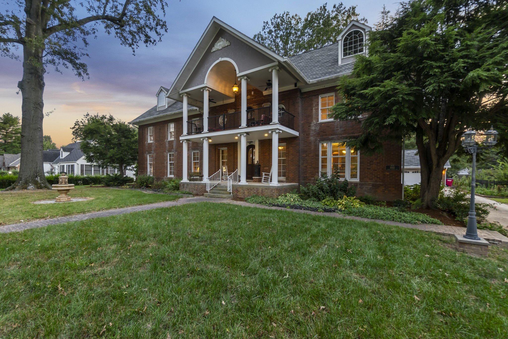 A large two-story brick house with a front porch and balcony, surrounded by a well-maintained lawn with trees, street lamps, and a walkway.