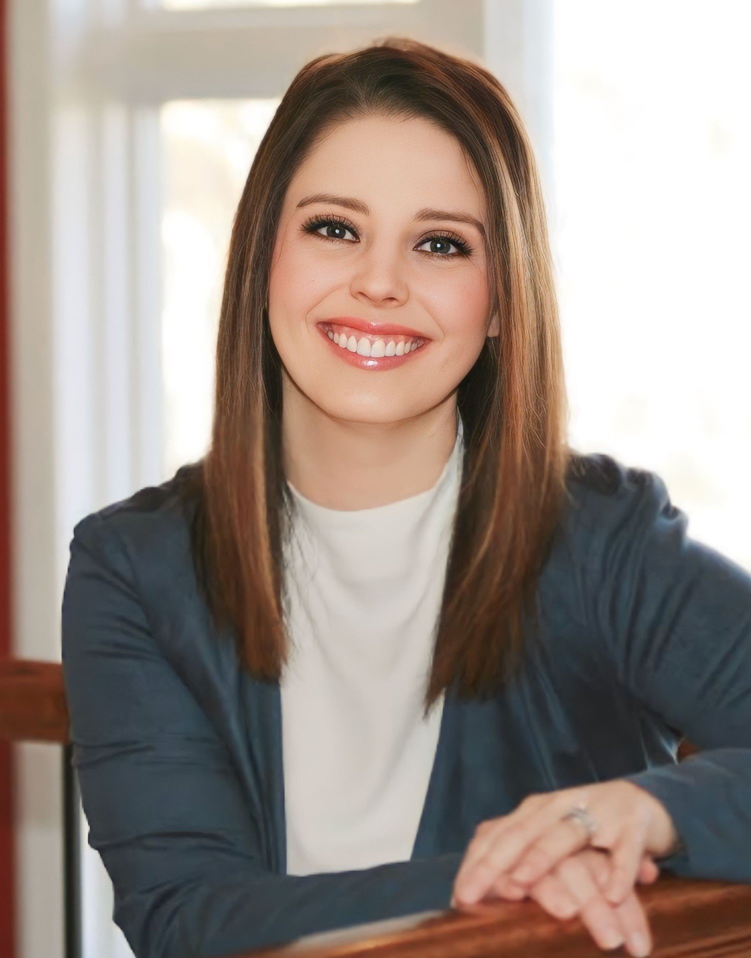 A young woman with brown hair and a white shirt, smiling, sitting at a wooden table with sunlight in the background.
